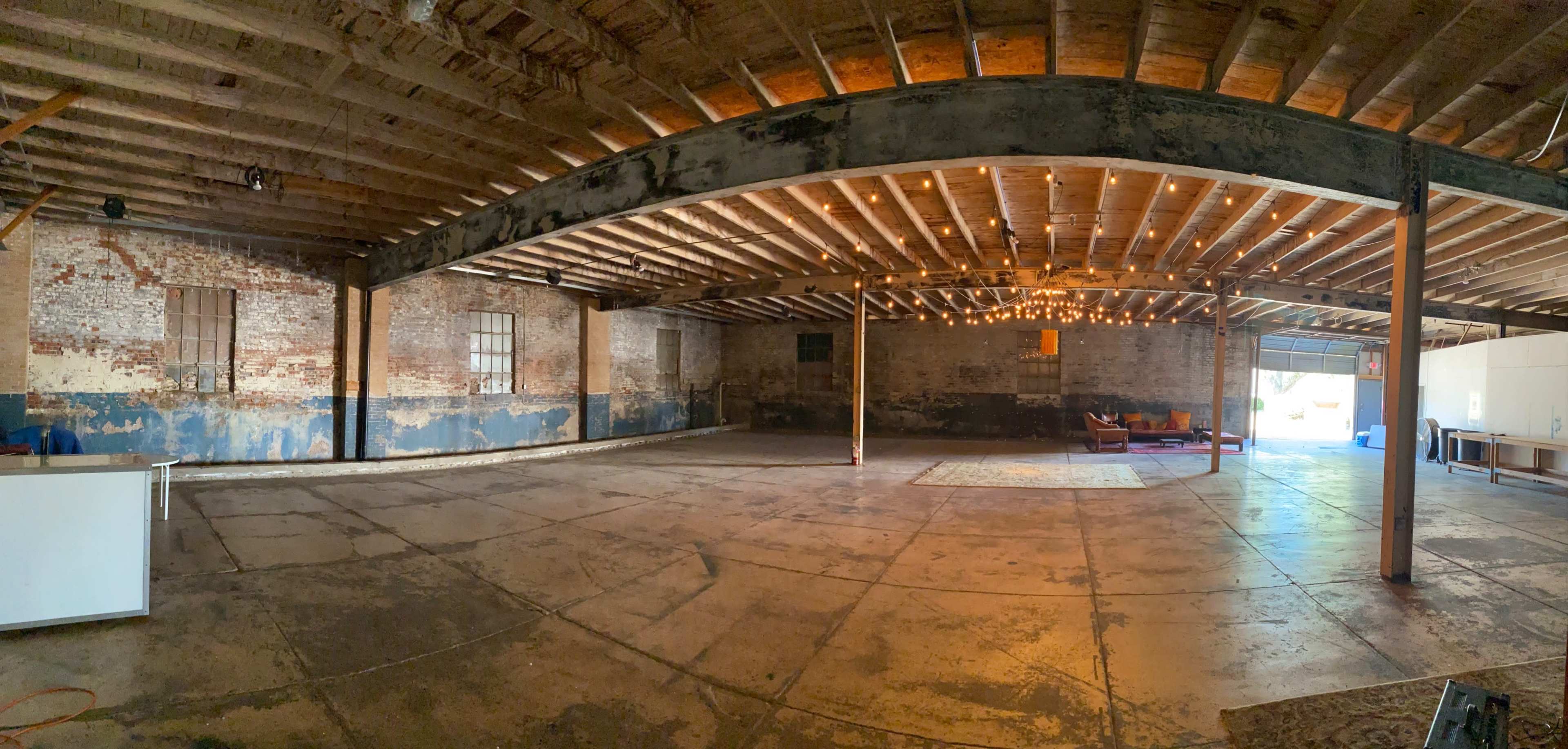 The image shows a spacious, empty industrial room with exposed brick walls, wooden beams, and large windows, featuring a few furniture pieces and an area rug.