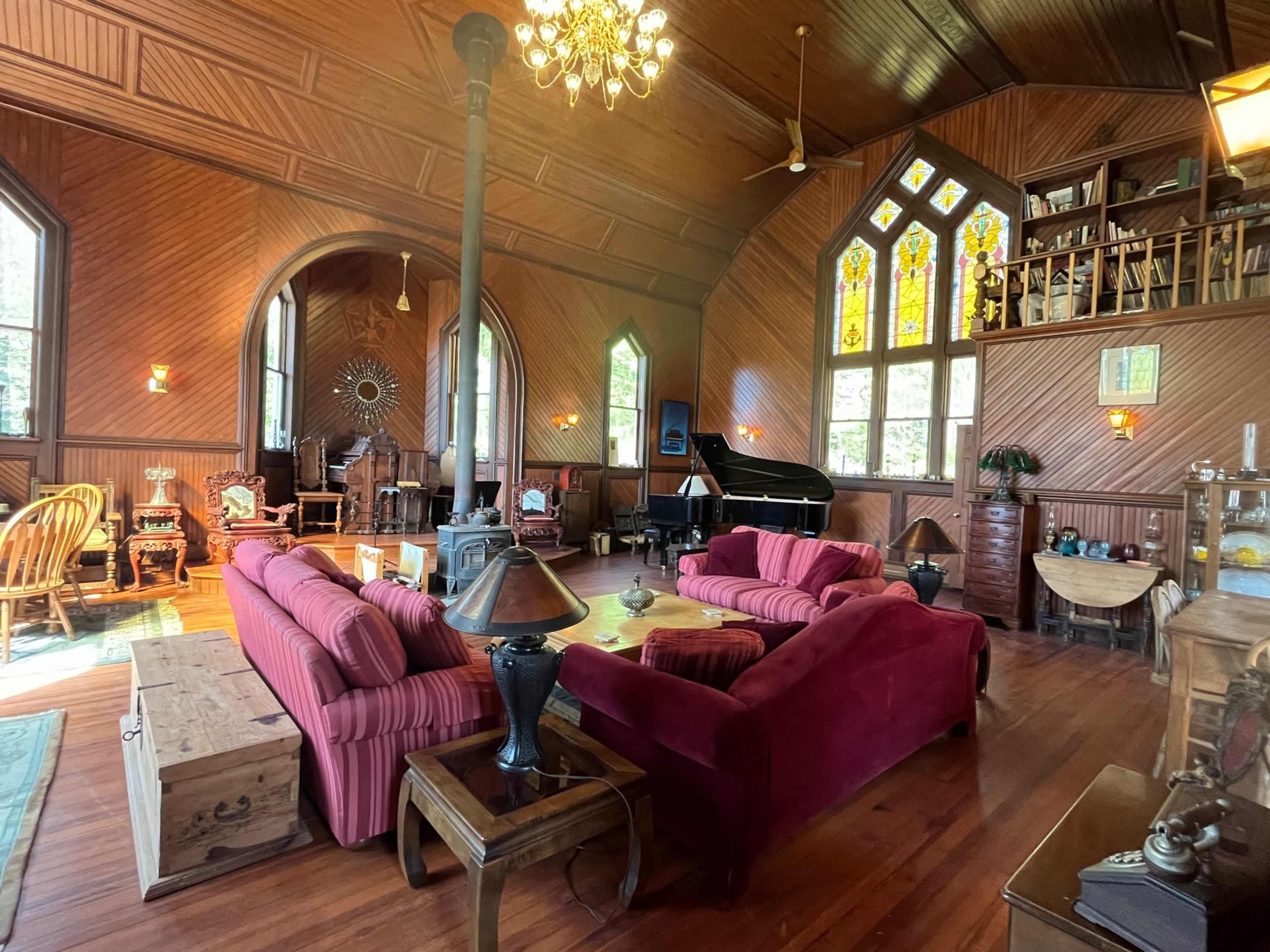 The room features a large, wood-paneled space with a grand piano, stained glass windows, and a mix of vintage furniture arranged around a central seating area.