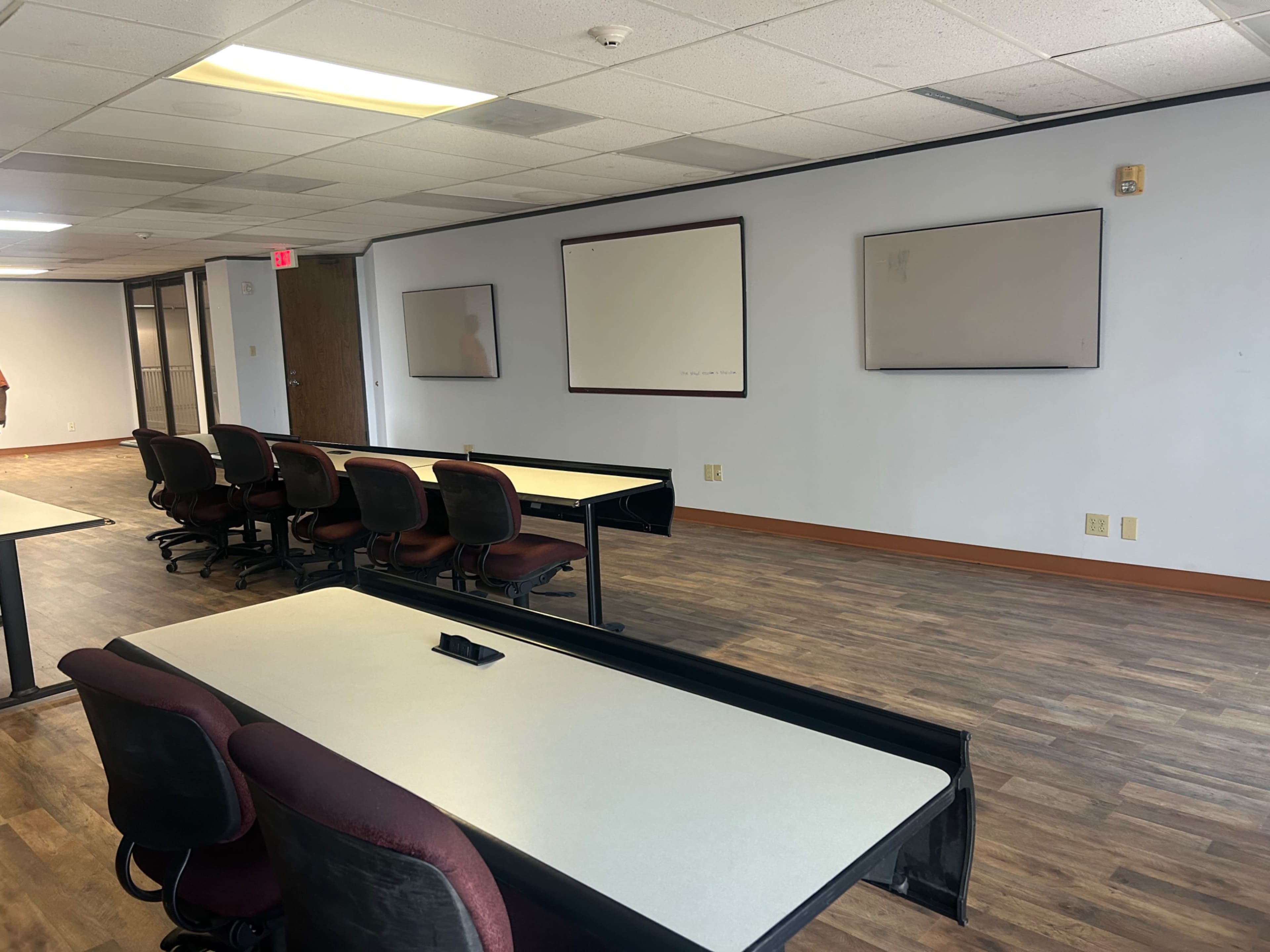 The image shows a conference room with several tables and chairs arranged facing blank whiteboards on the walls.
