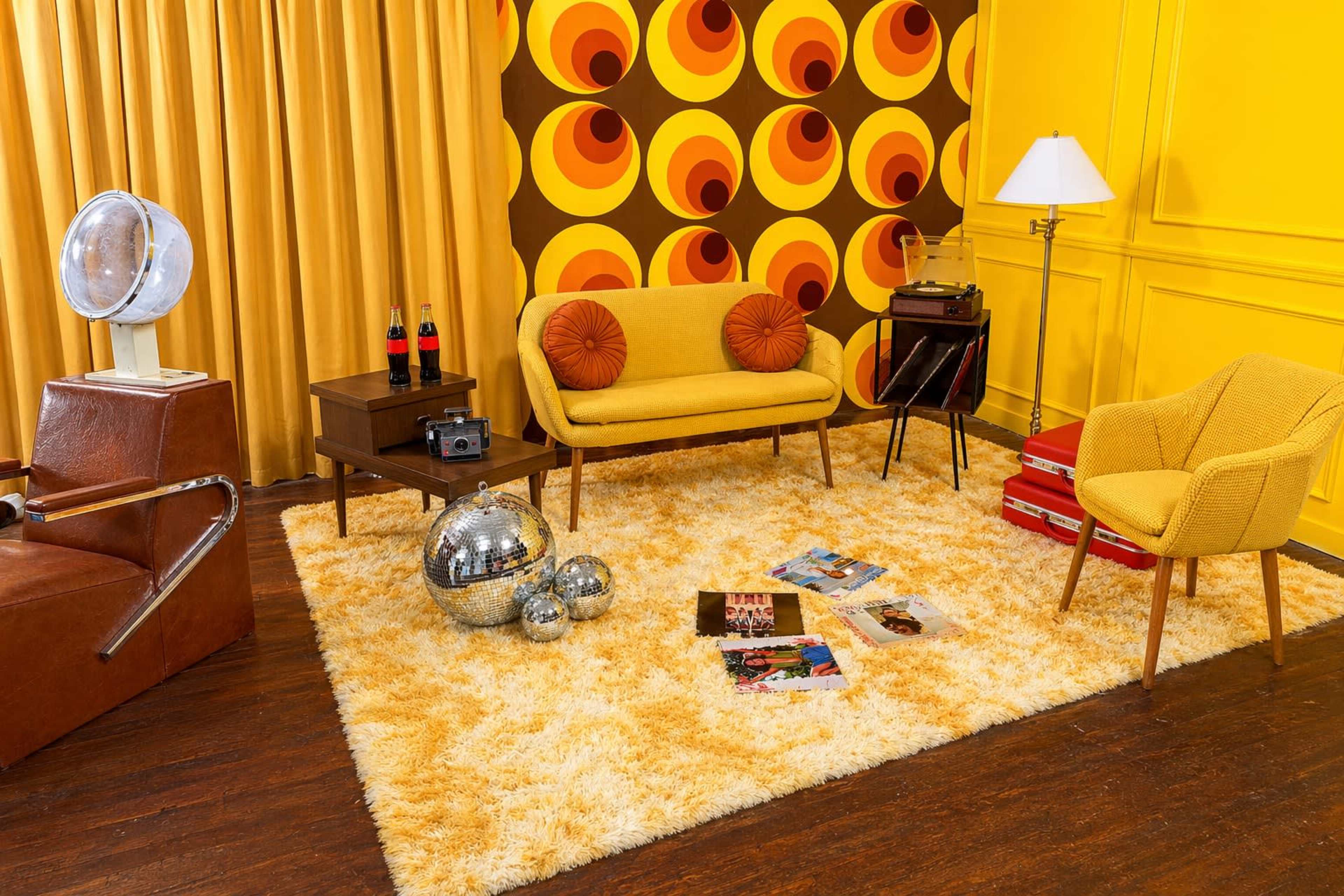 The image features a retro-inspired living room with yellow walls, a patterned backdrop, vintage furniture, and decorative items like a disco ball and vinyl records.