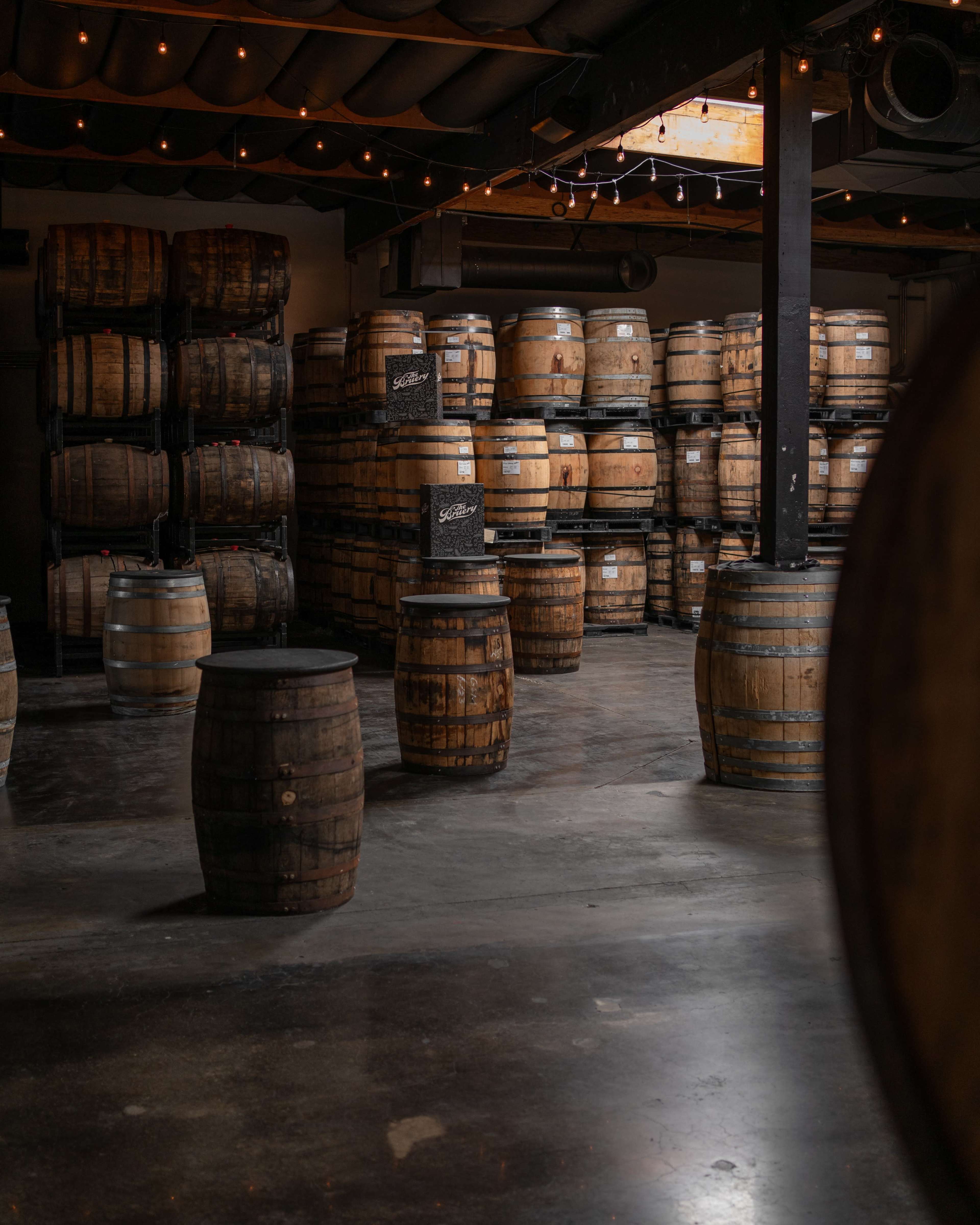 The image shows a dimly lit warehouse filled with stacked wooden barrels arranged neatly against the walls and several standing barrels in the foreground.