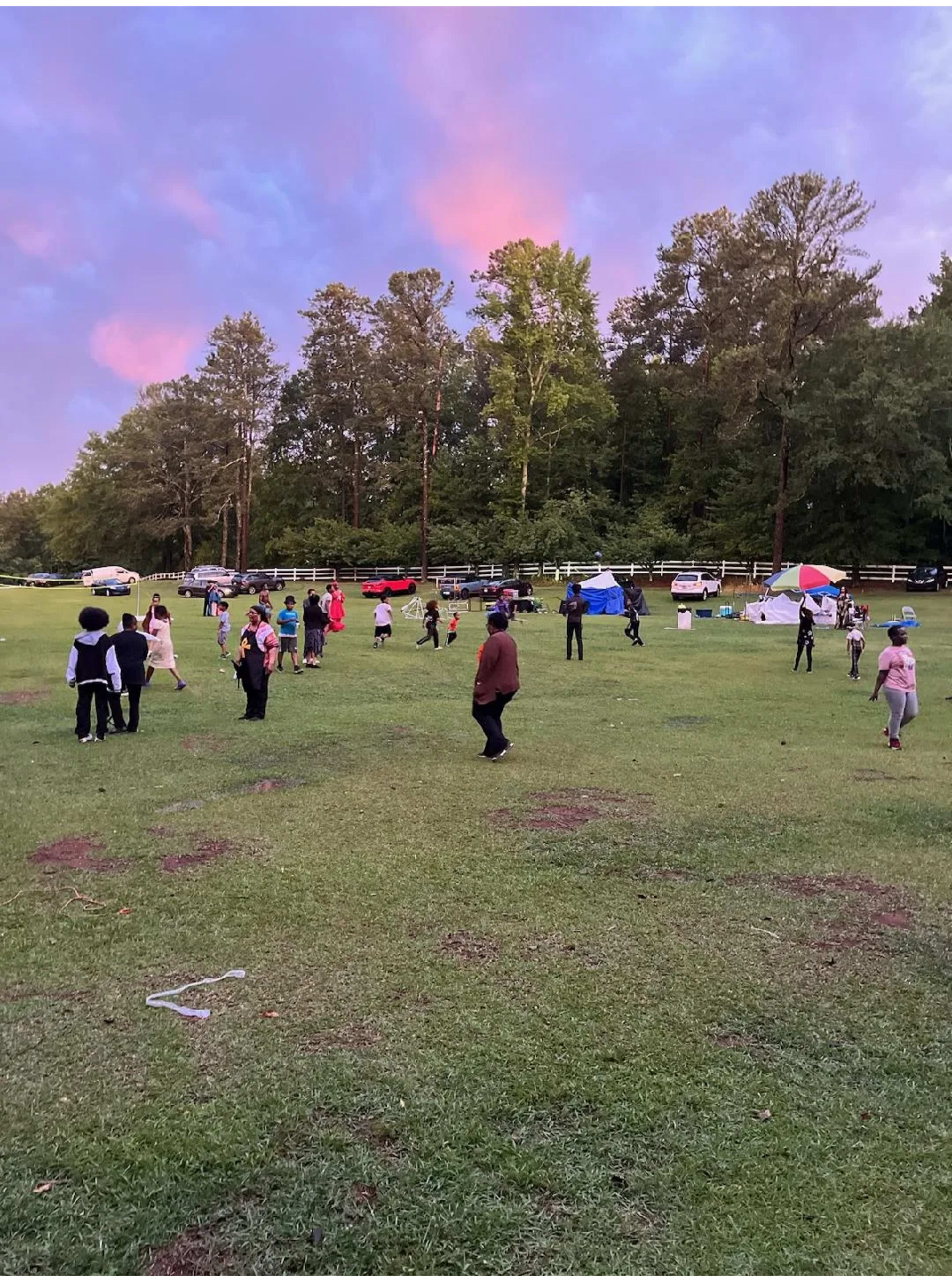 A group of people are gathered on a grassy field, some engaging in activities while others are scattered throughout the area, with cars and tents visible in the background.