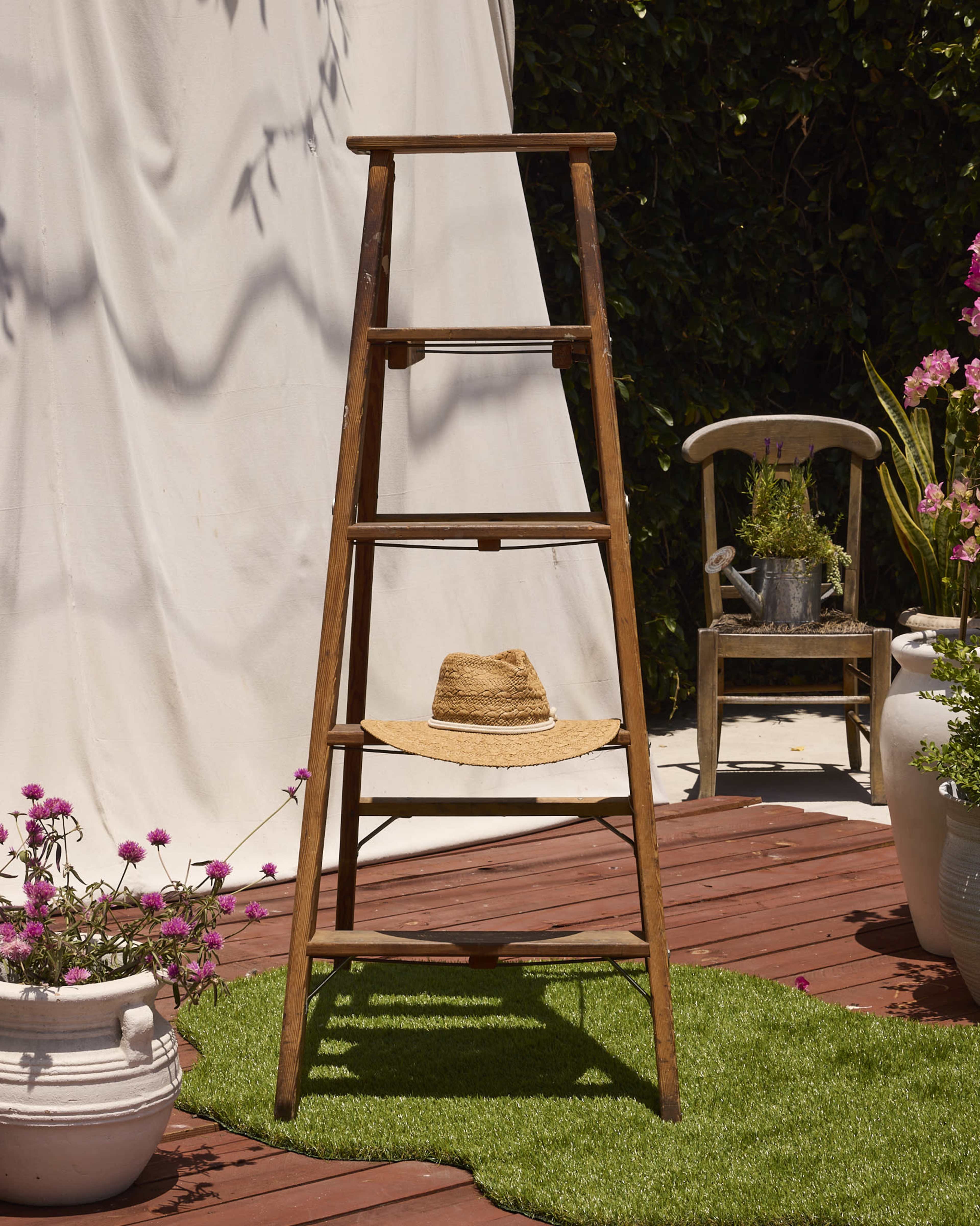 A wooden three-step ladder stands on a wooden deck, with a straw hat resting on its second step, surrounded by potted flowers and a chair in the background.