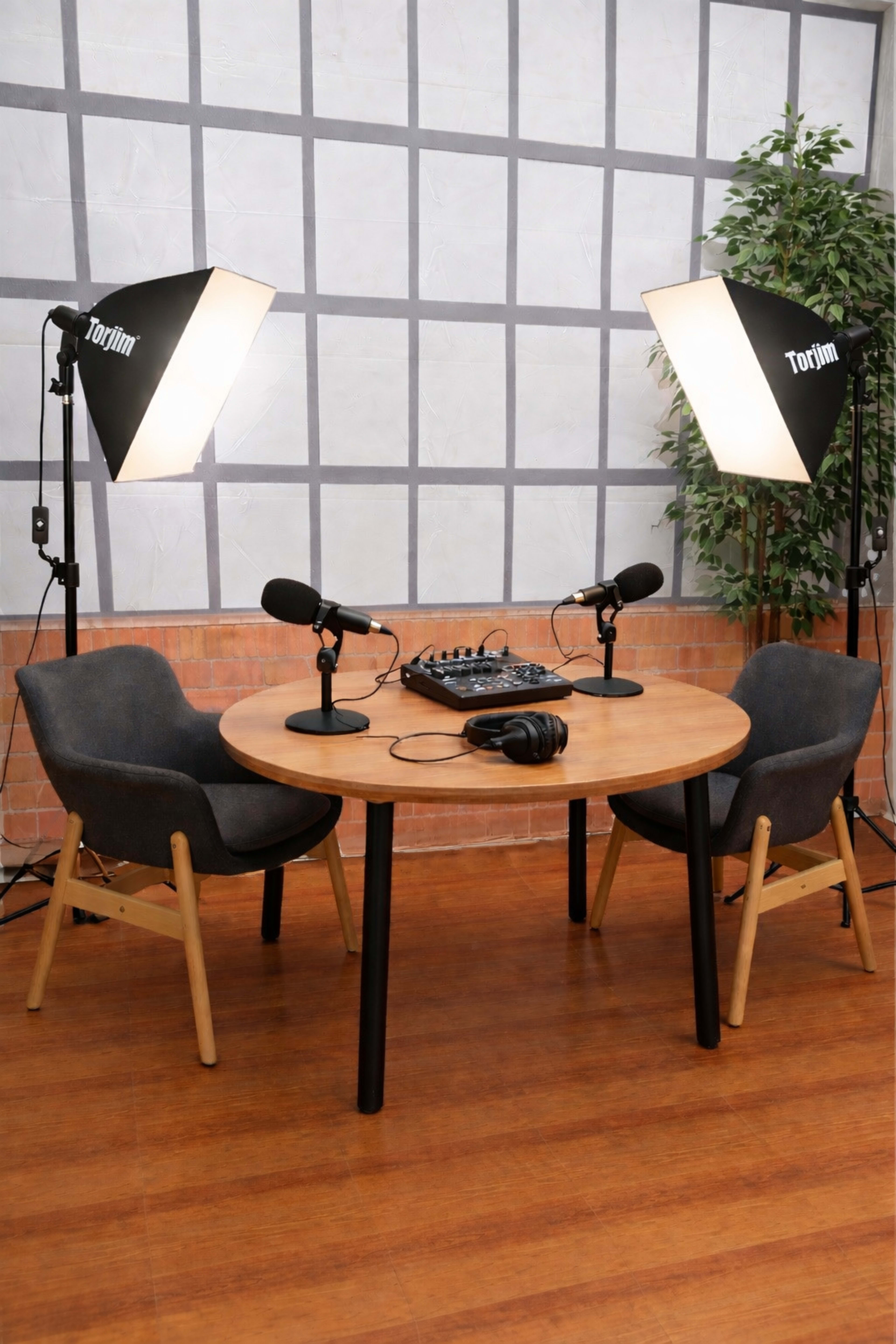 A circular wooden table with two microphones, headphones, and audio equipment is set between two chairs under studio lighting in a sound recording space.