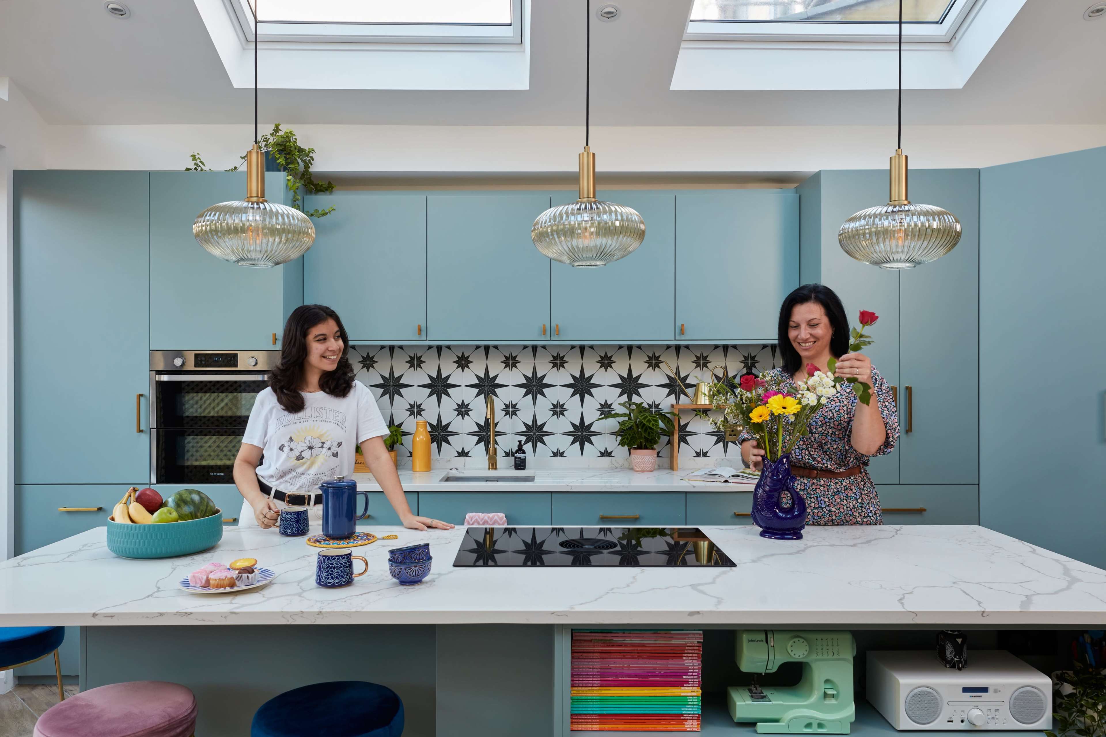 Two women are arranging flowers in a modern kitchen with blue cabinets and patterned tiles, while items like fruit and dishes are displayed on the island.