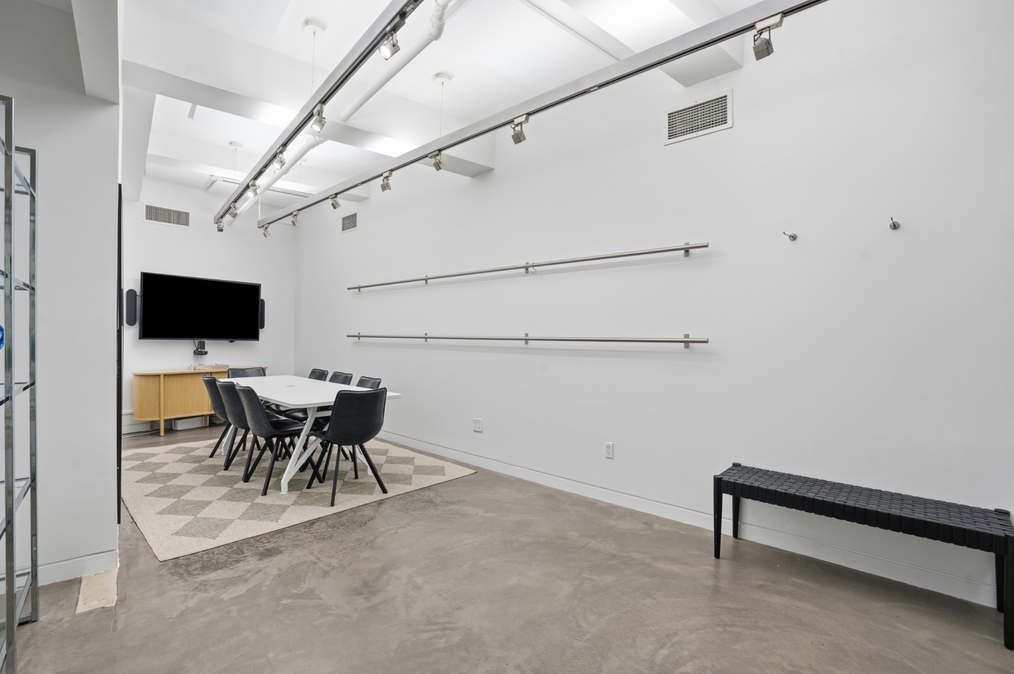 The image shows a modern, minimalistic meeting room with a long table, black chairs, and wall-mounted shelves, set against a white background and with polished concrete flooring.