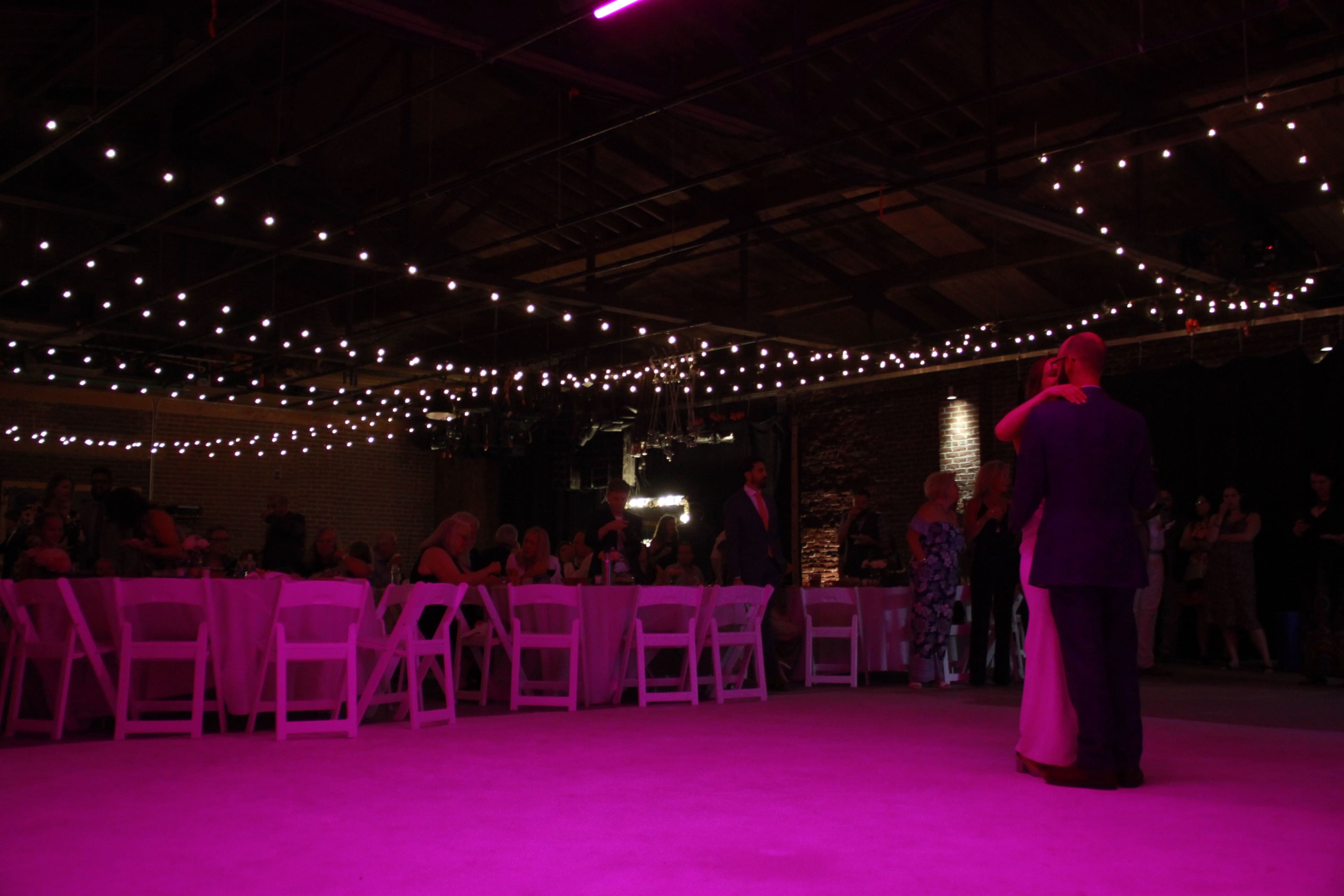 A couple dances on a lighted floor beneath strings of lights in a large indoor event space while guests sit at round tables.