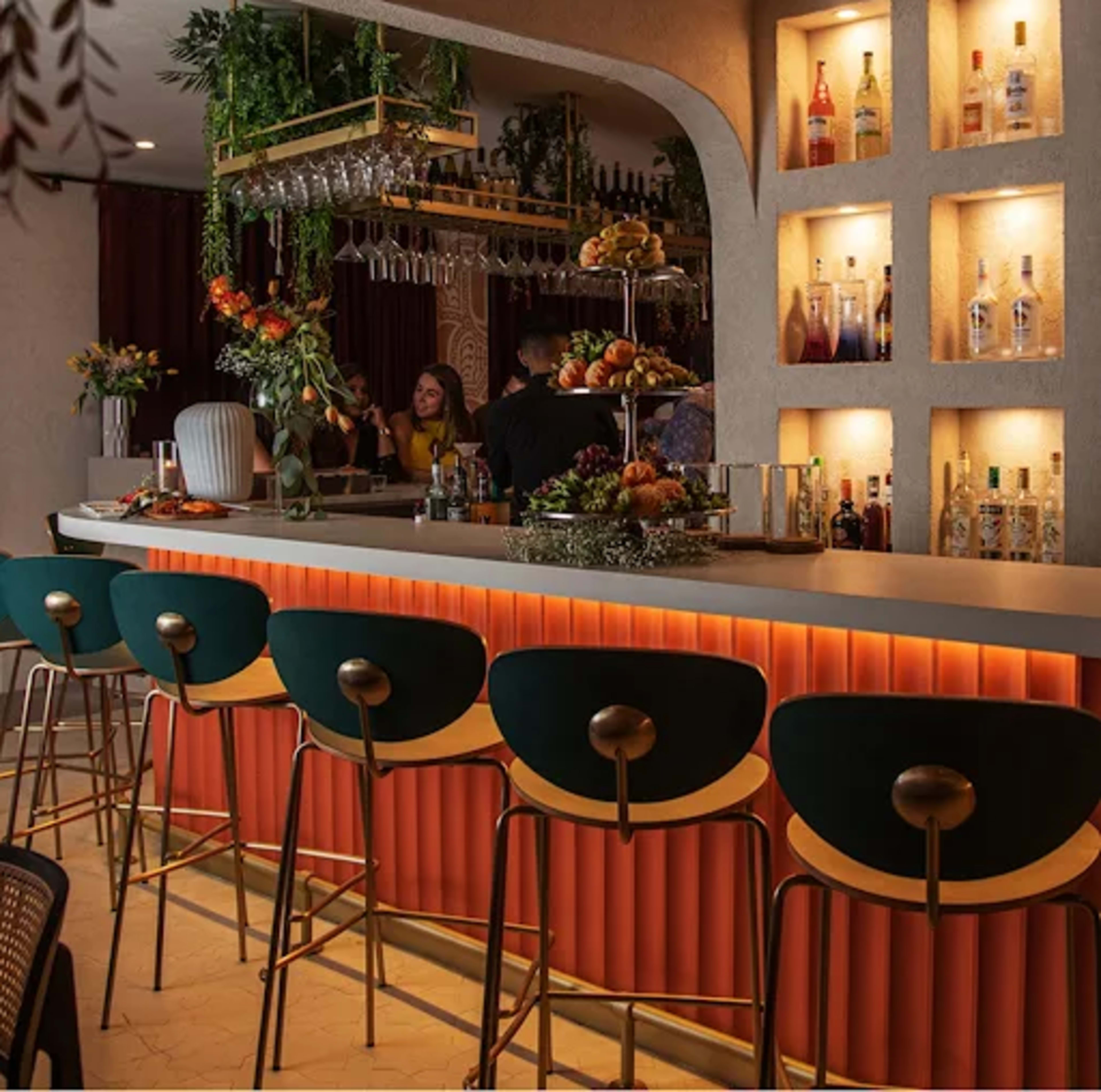 The image shows a stylish bar area featuring a long counter with curved stools, shelves displaying a variety of bottles, and decorative fruits on a tiered stand.