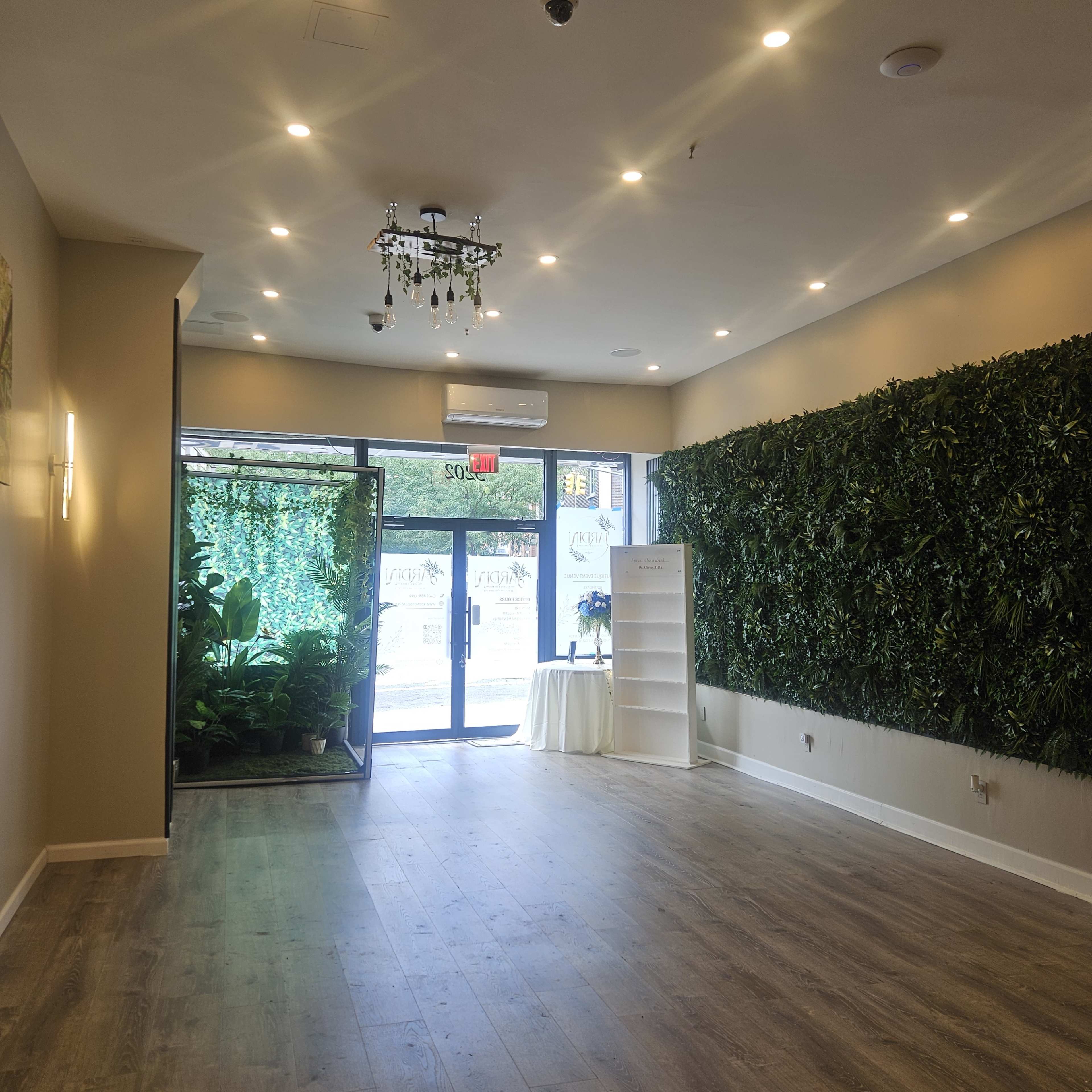 The image shows an empty room with a wooden floor, featuring a green wall of plants on one side and a glass door leading outside.