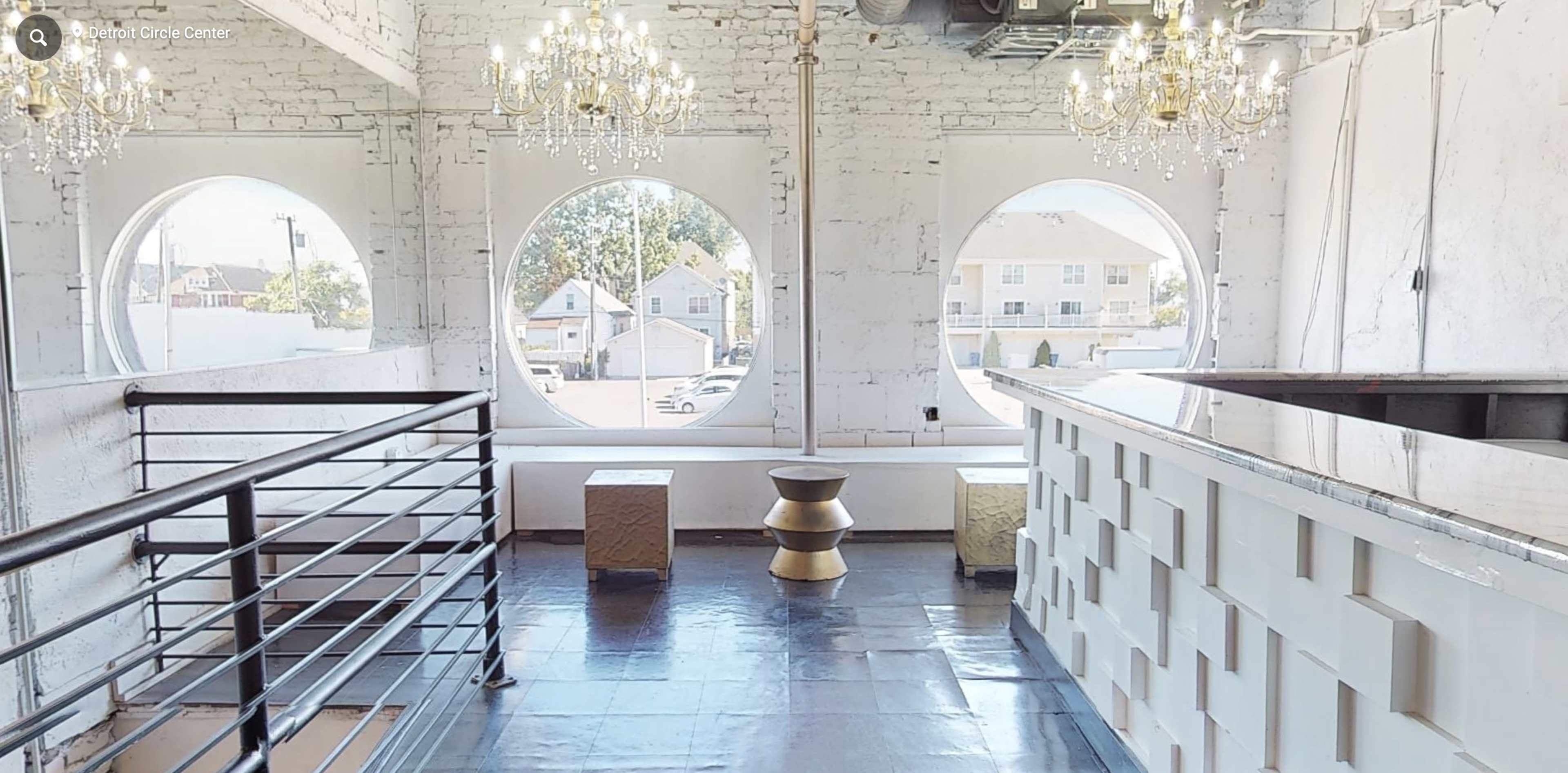 The interior space features large circular windows, two hanging chandeliers, and a minimalist design with a bar area and metallic tables.