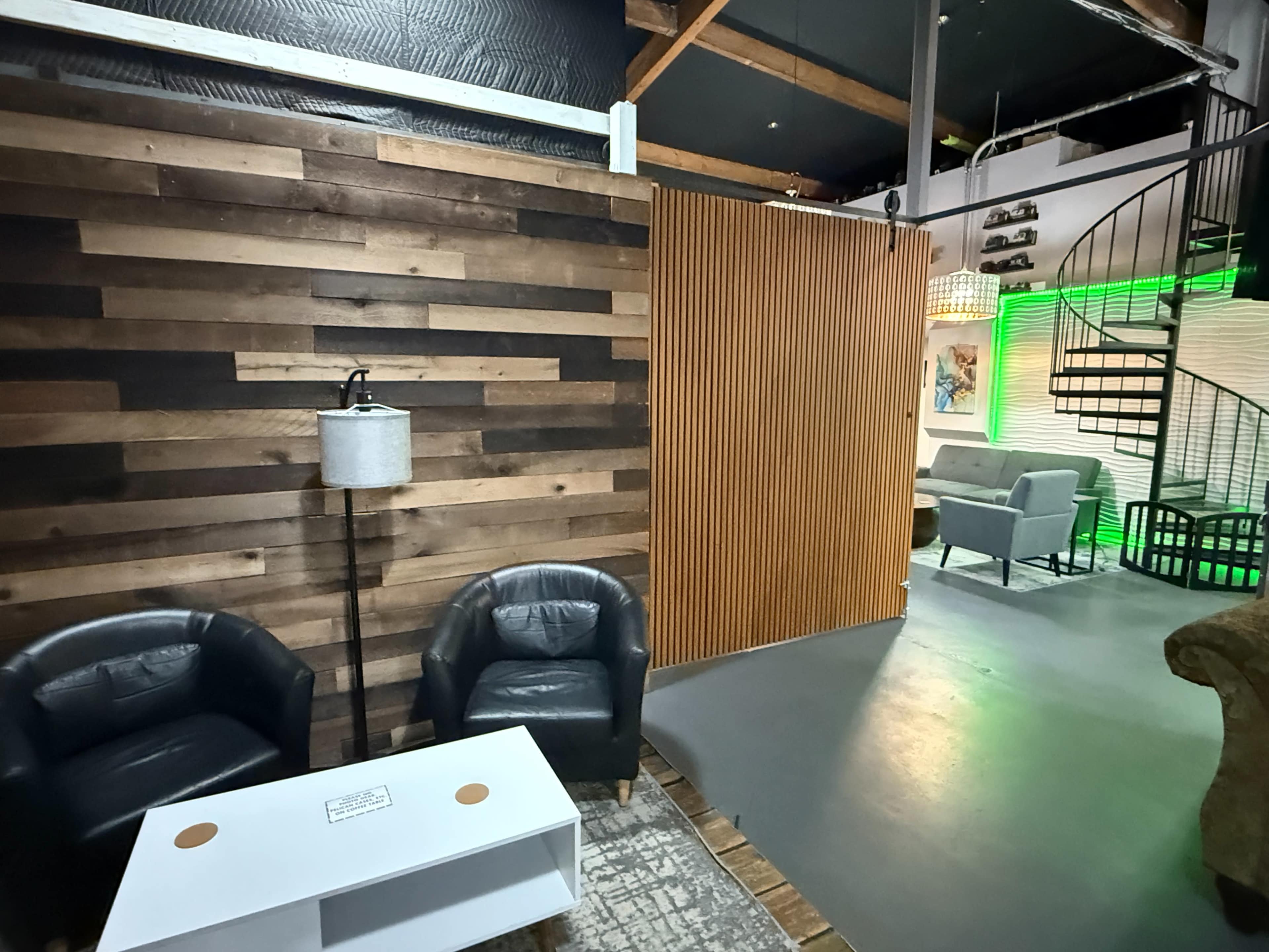 The image shows a modern interior space featuring wooden accent walls, two black leather chairs, a small white coffee table, and a spiral staircase in the background.