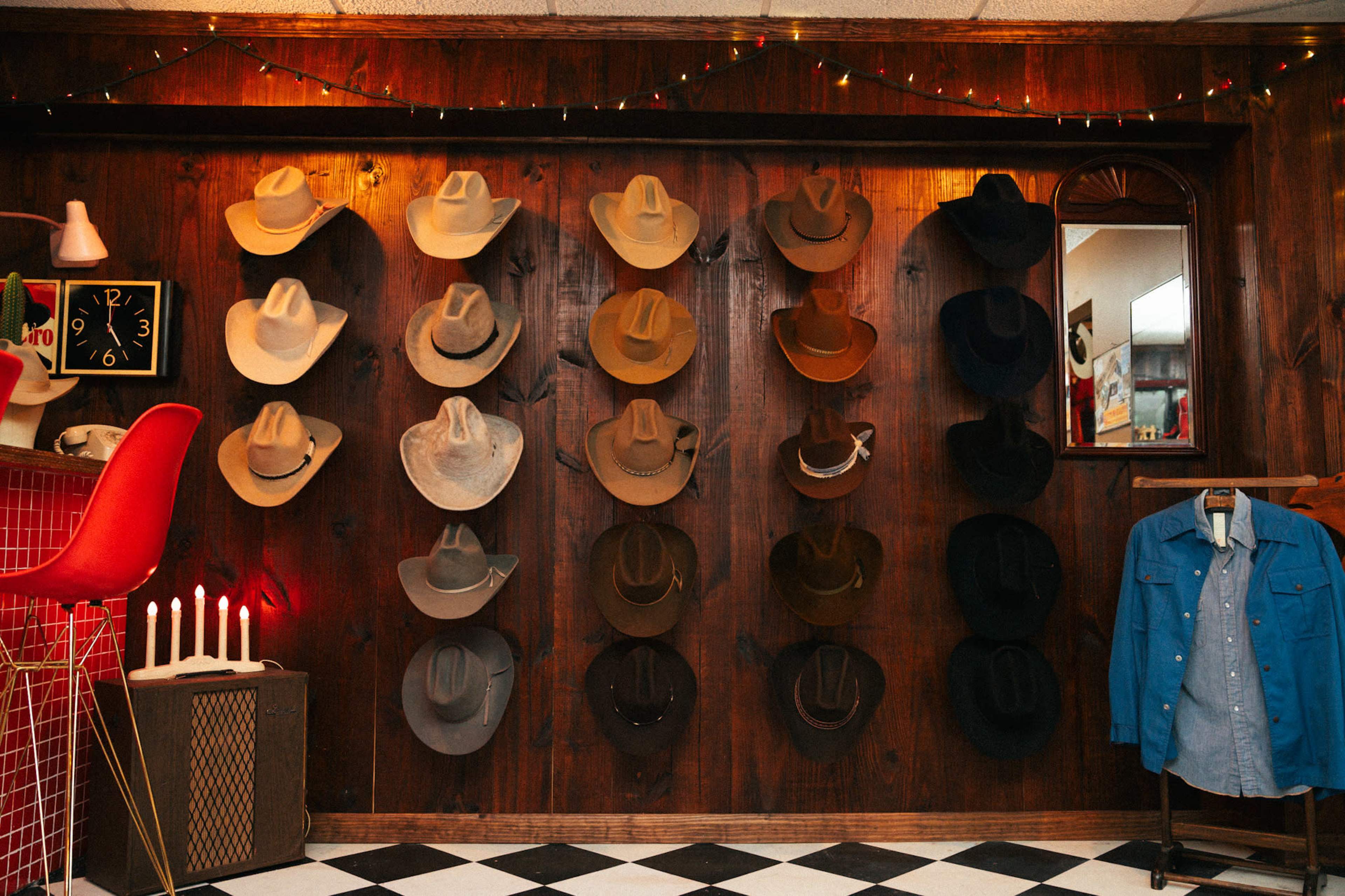 The image shows a wall adorned with various cowboy hats hanging alongside a mirror, with a red and black checkerboard floor and a blue shirt displayed on a hanger.