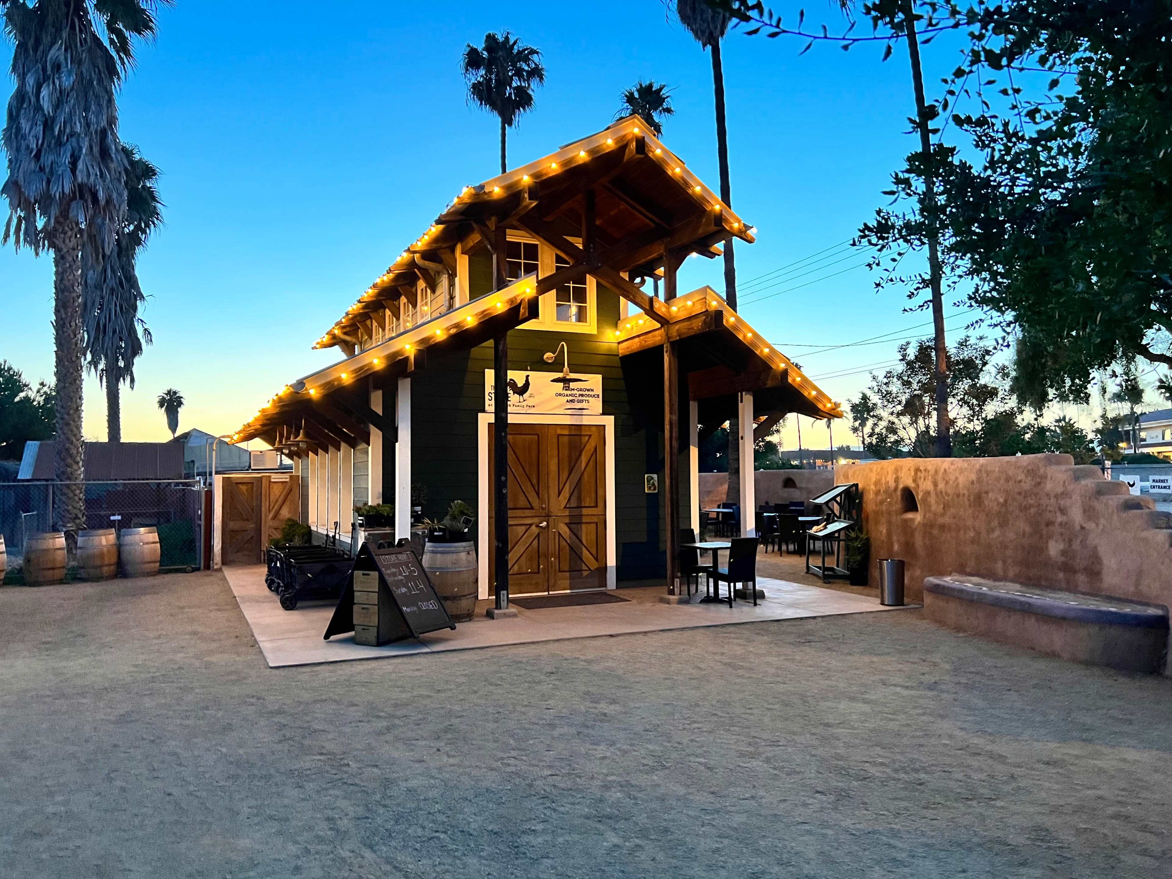 A two-story building with a modern rustic design, featuring outdoor seating and decorative string lights, surrounded by palm trees at dusk.