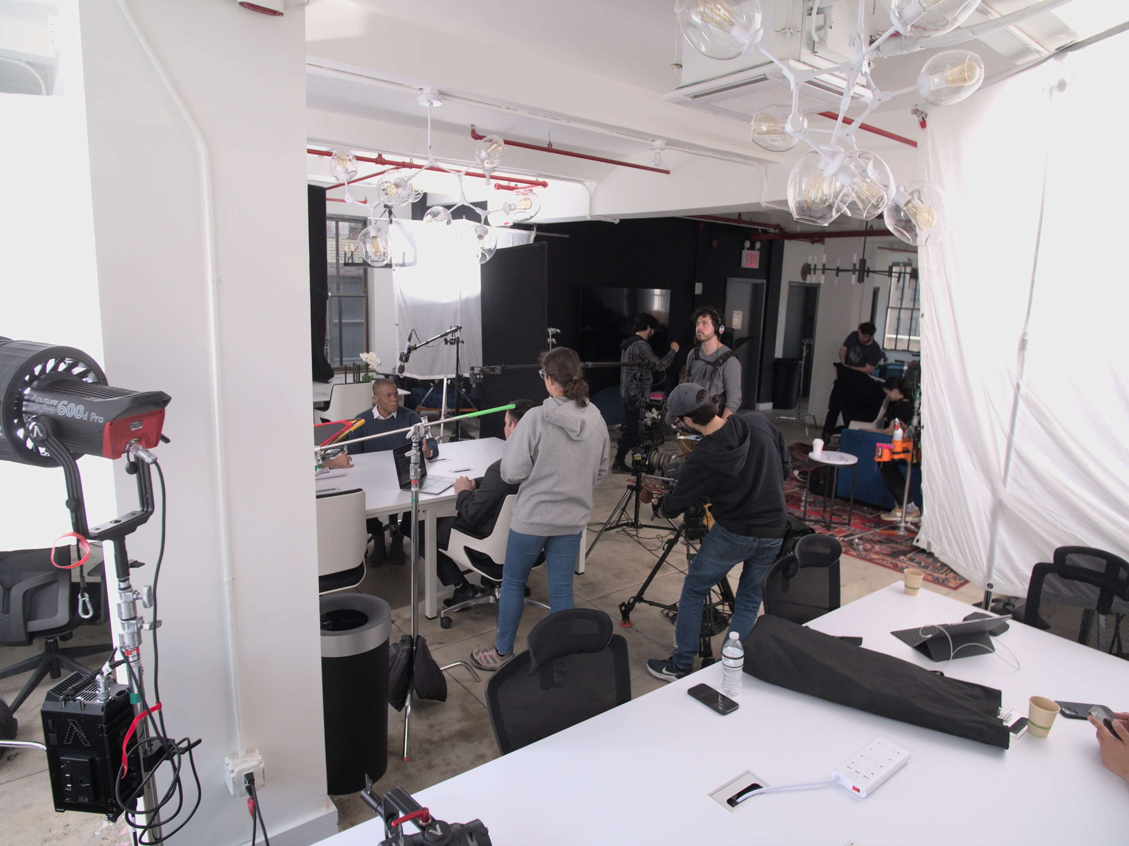 A group of people is filming a scene in a bright, modern workspace equipped with cameras and lighting.