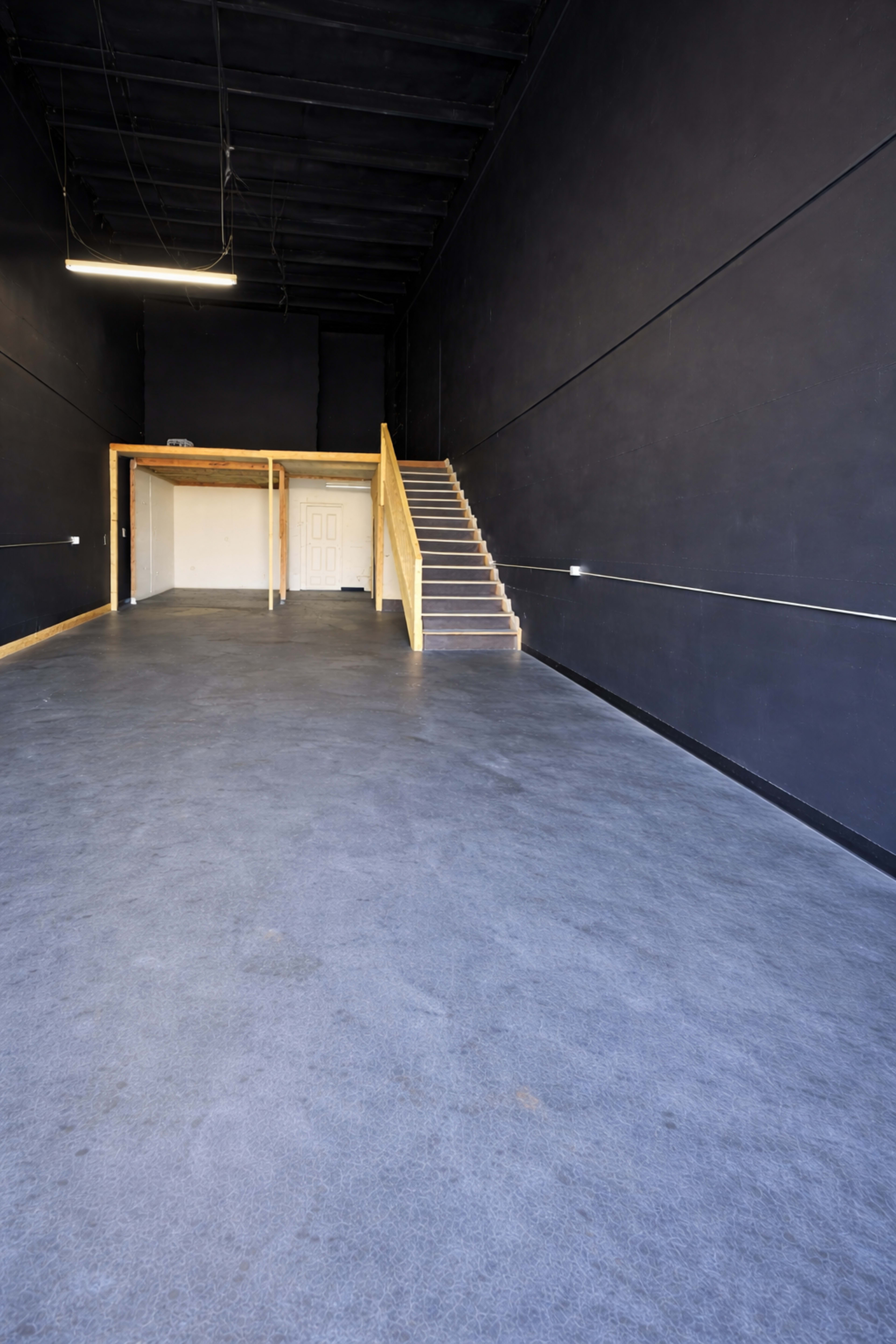The image shows an empty, spacious room with dark walls, a concrete floor, and a staircase leading to a second level.