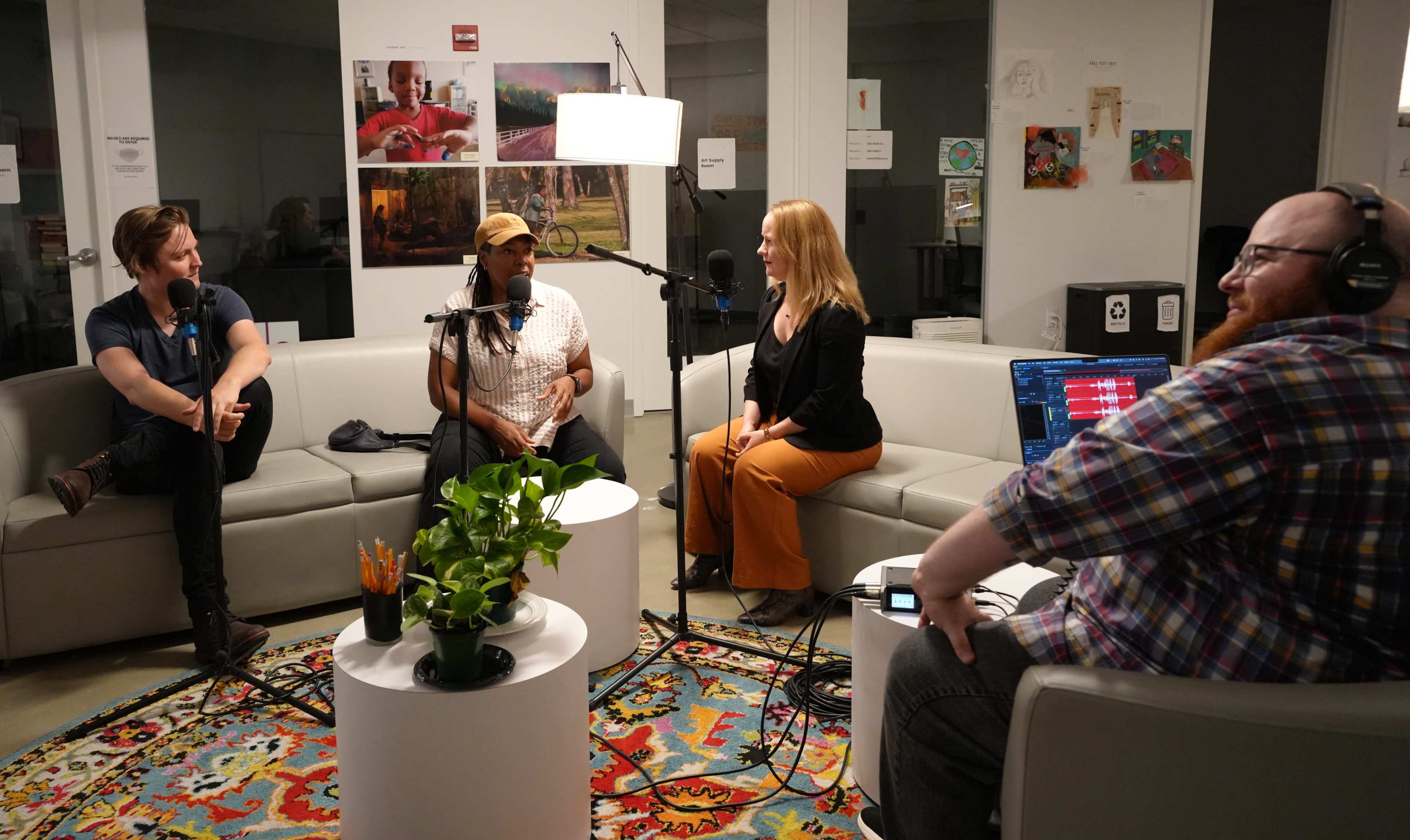 Four individuals participate in a discussion in a modern lounge area with a microphone and recording equipment set up on a round table.