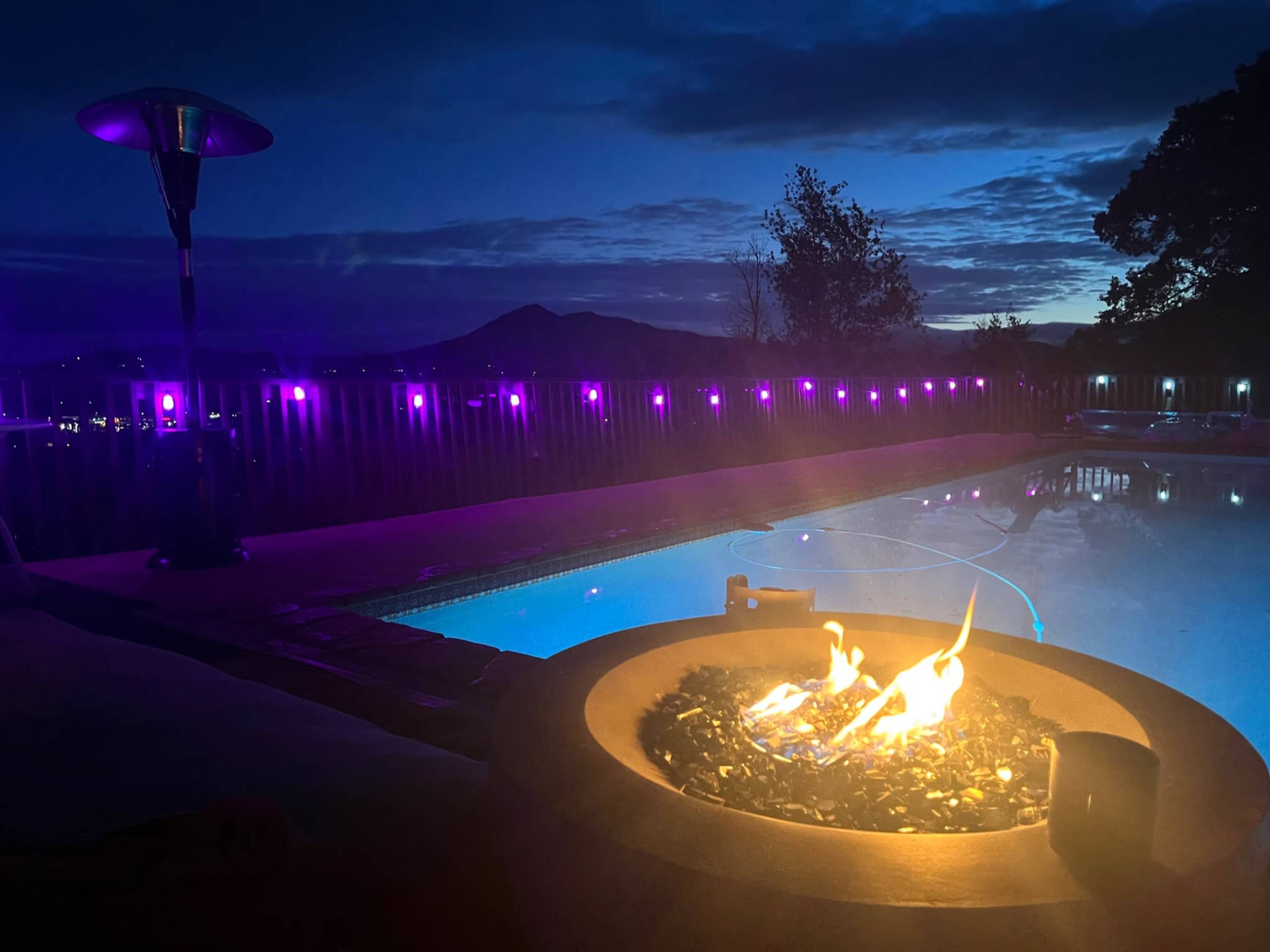 A poolside fire pit illuminates the scene with flames, while purple lights outline the pool area against a twilight sky.