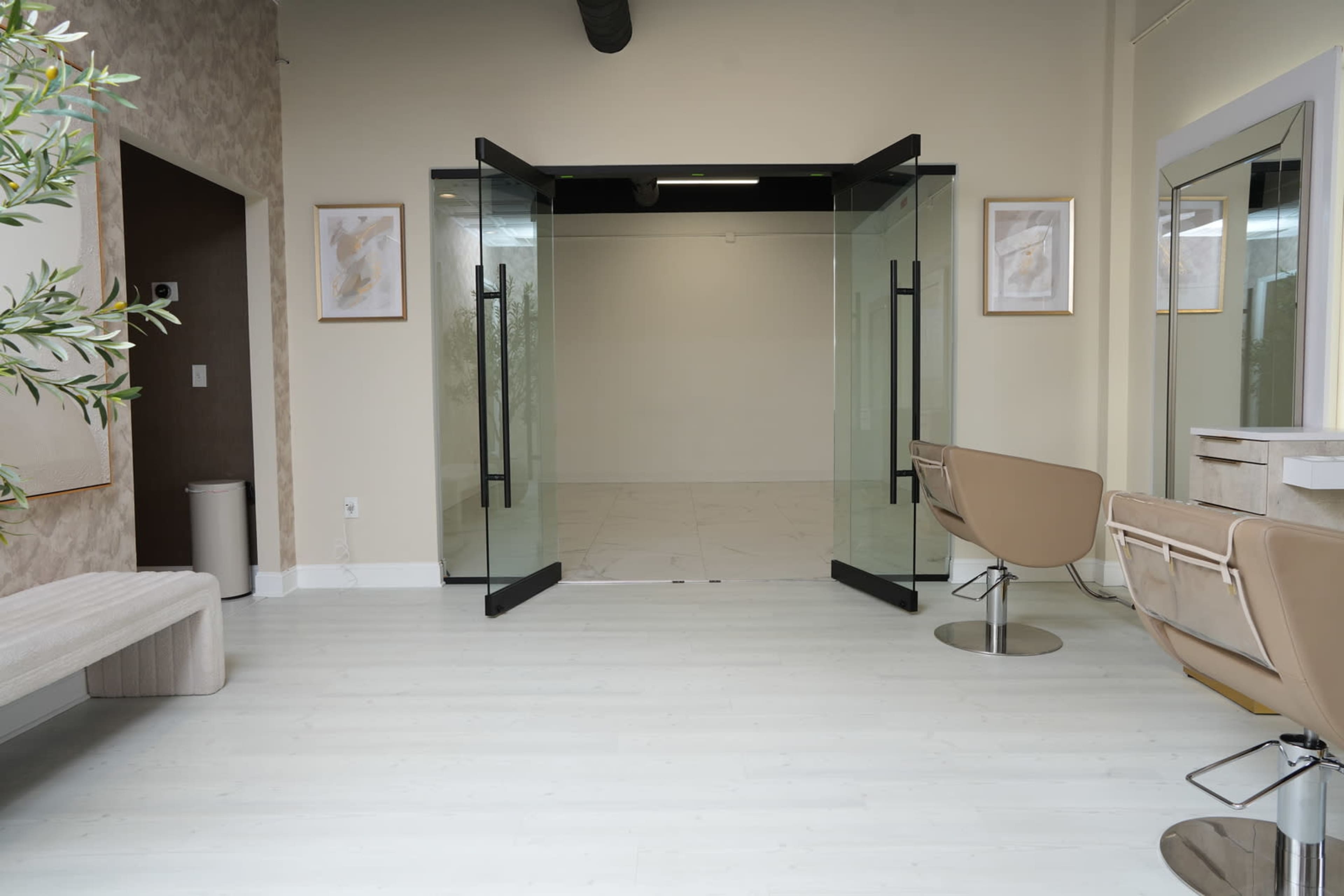 A modern salon interior features glass doors that open to a spacious reception area with minimalist decor and two stylist chairs.