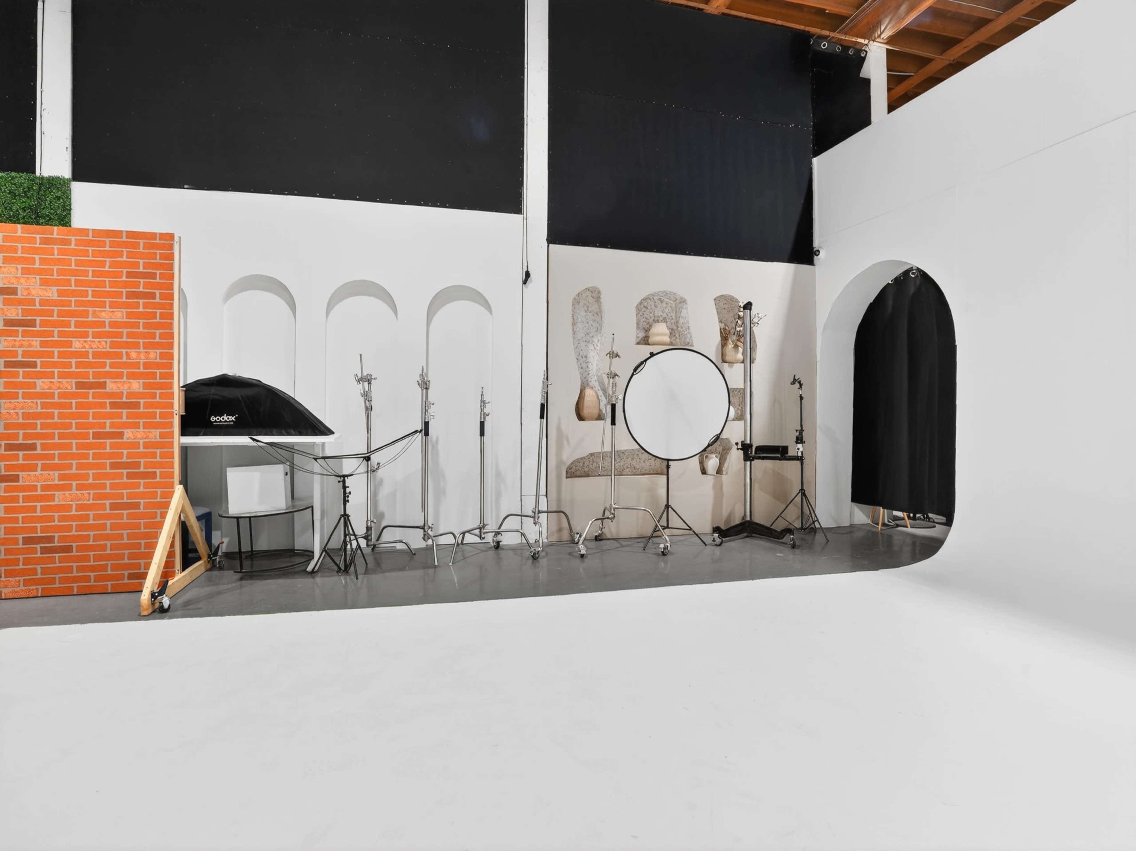 The image shows a photography studio with a white backdrop, a standing mirror, photography equipment, and a brick wall section on one side.