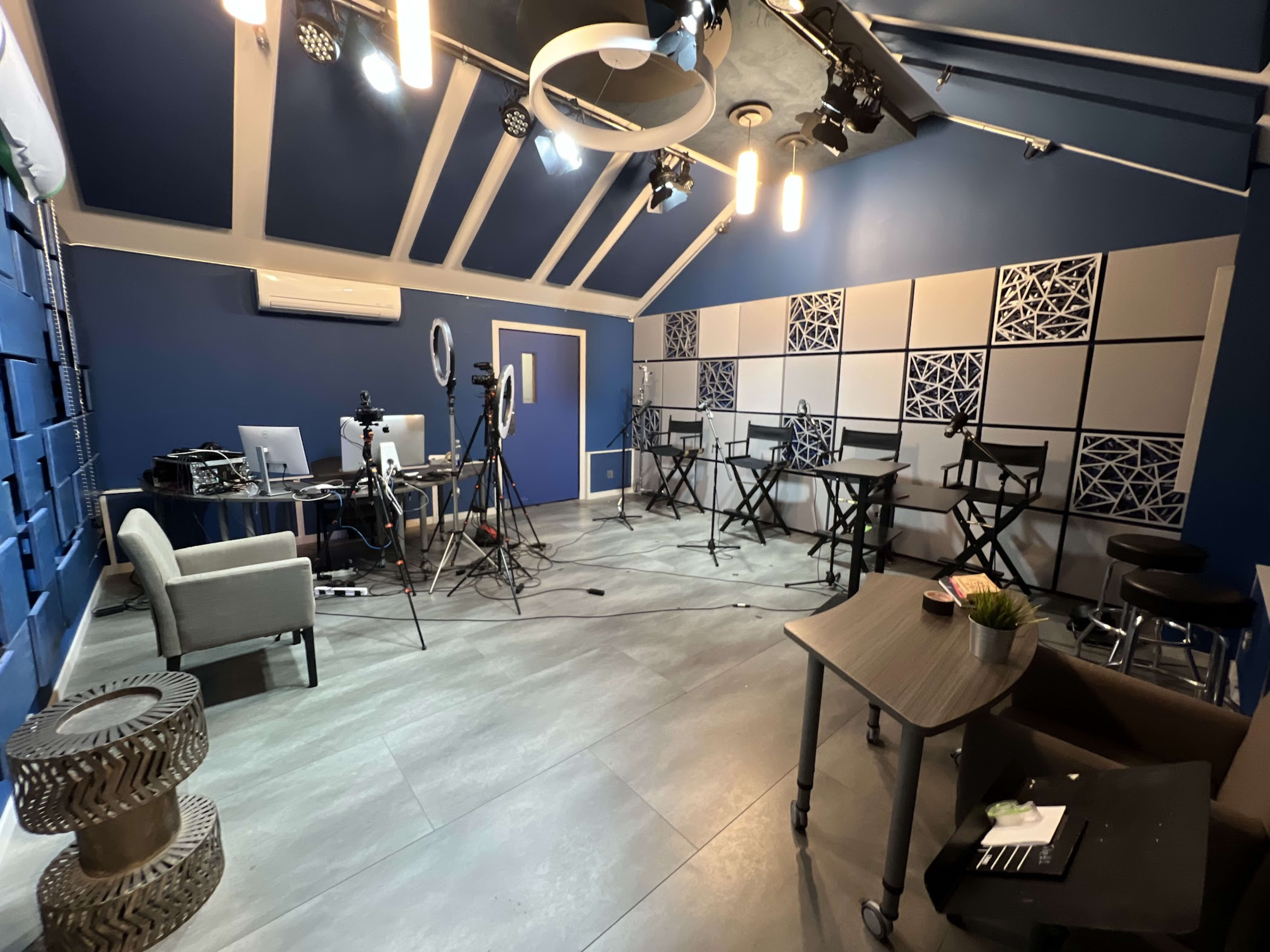 A modern studio setup with a blue wall, lighting fixtures, chairs, a table, and equipment for video recording.