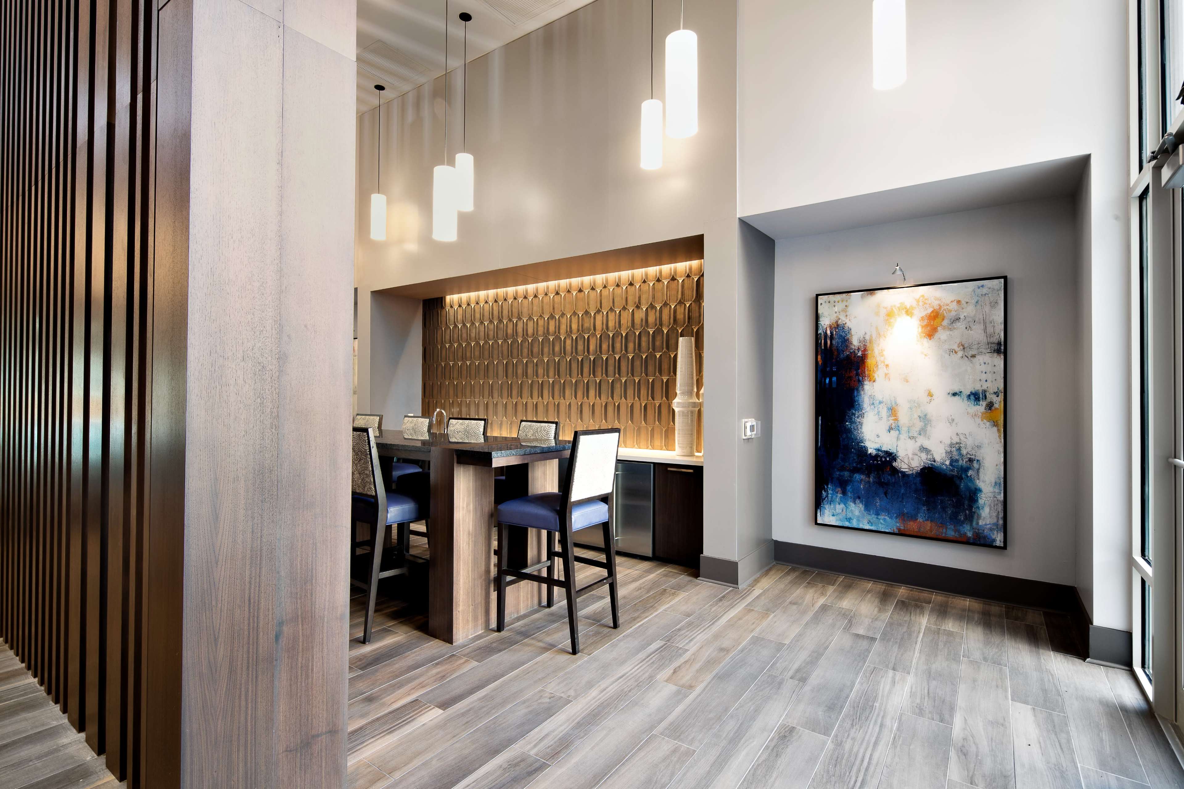 A modern interior space featuring a dining area with a high table and chairs, wooden paneling, and an abstract painting on the wall.