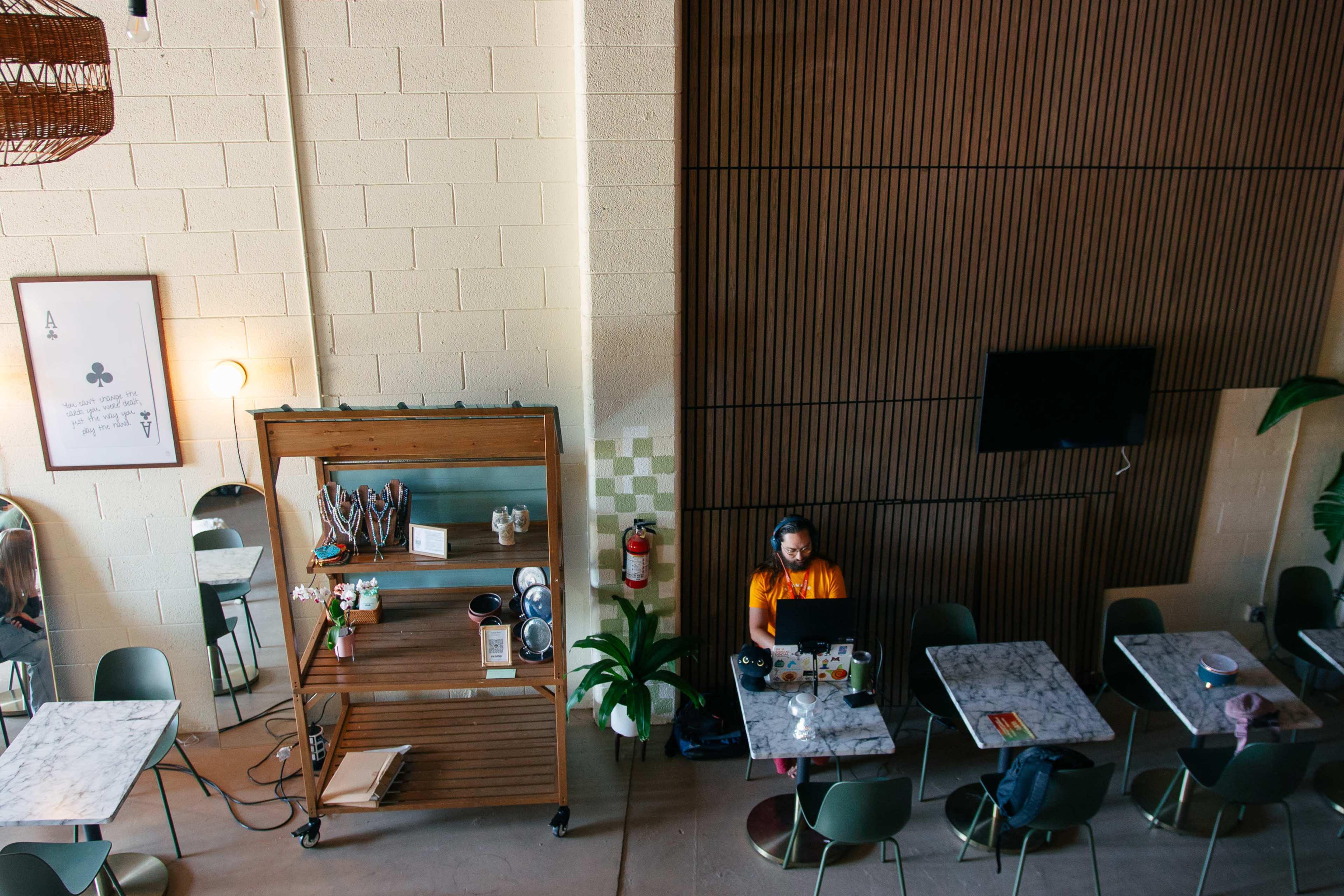 A person sits at a table in a café, working on a laptop with headphones on, while marble tables and a wooden feature wall are visible in the background.