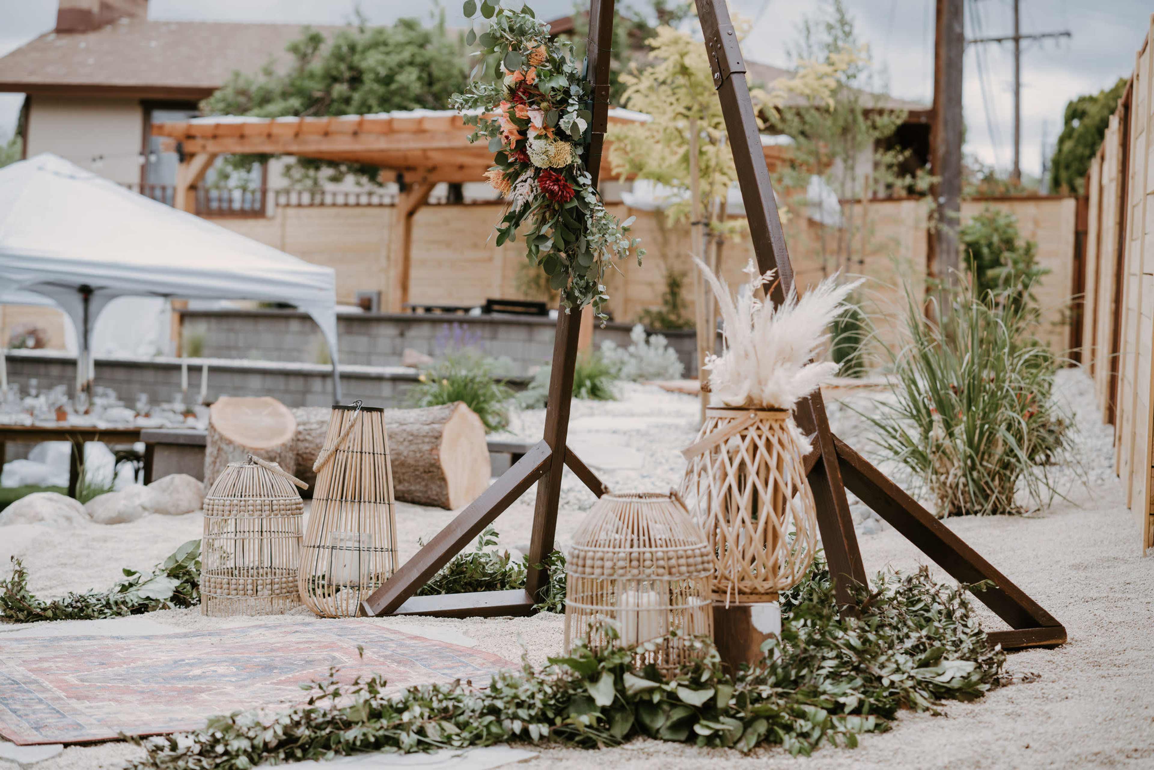 The scene features a decorated outdoor area with a wooden arch adorned with flowers, surrounded by lanterns and natural elements on sand.