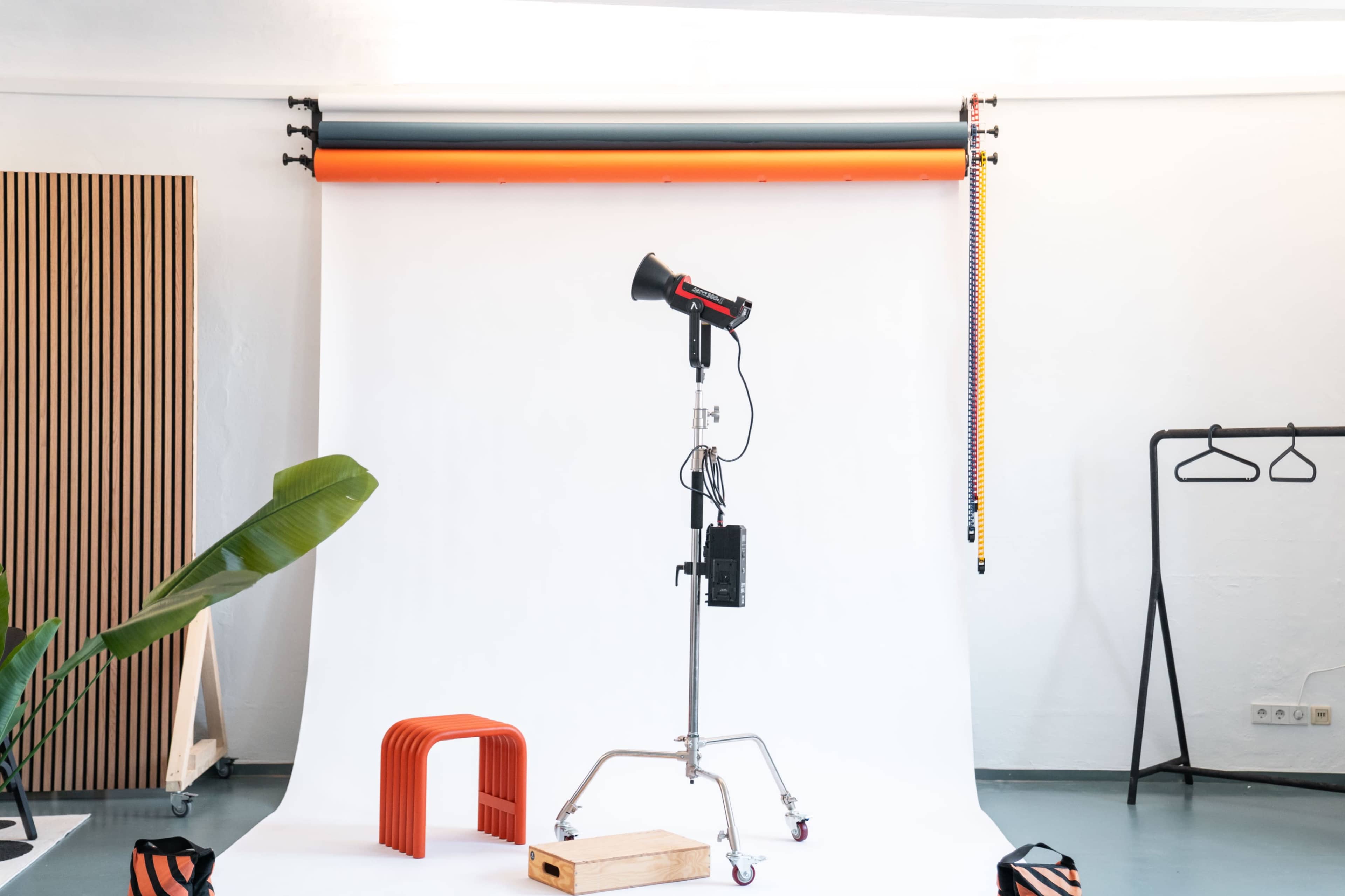 A photography studio setup features a backdrop, a spotlight on a stand, a wooden platform, and a few props, including an orange stool and a hanging rack.