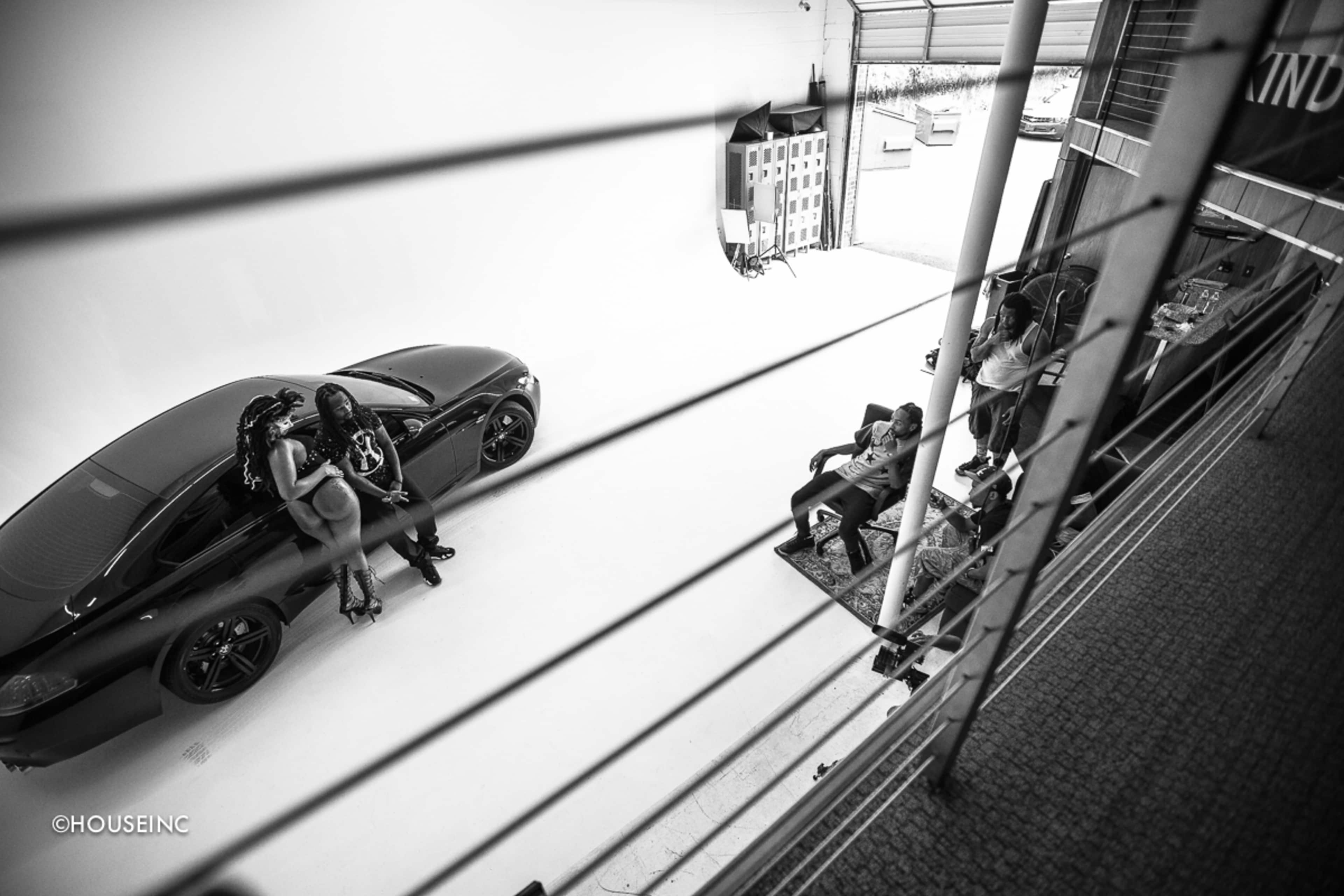 A large, modern photo studio features a black car and two women posing beside it, while two individuals sit on a rug nearby.