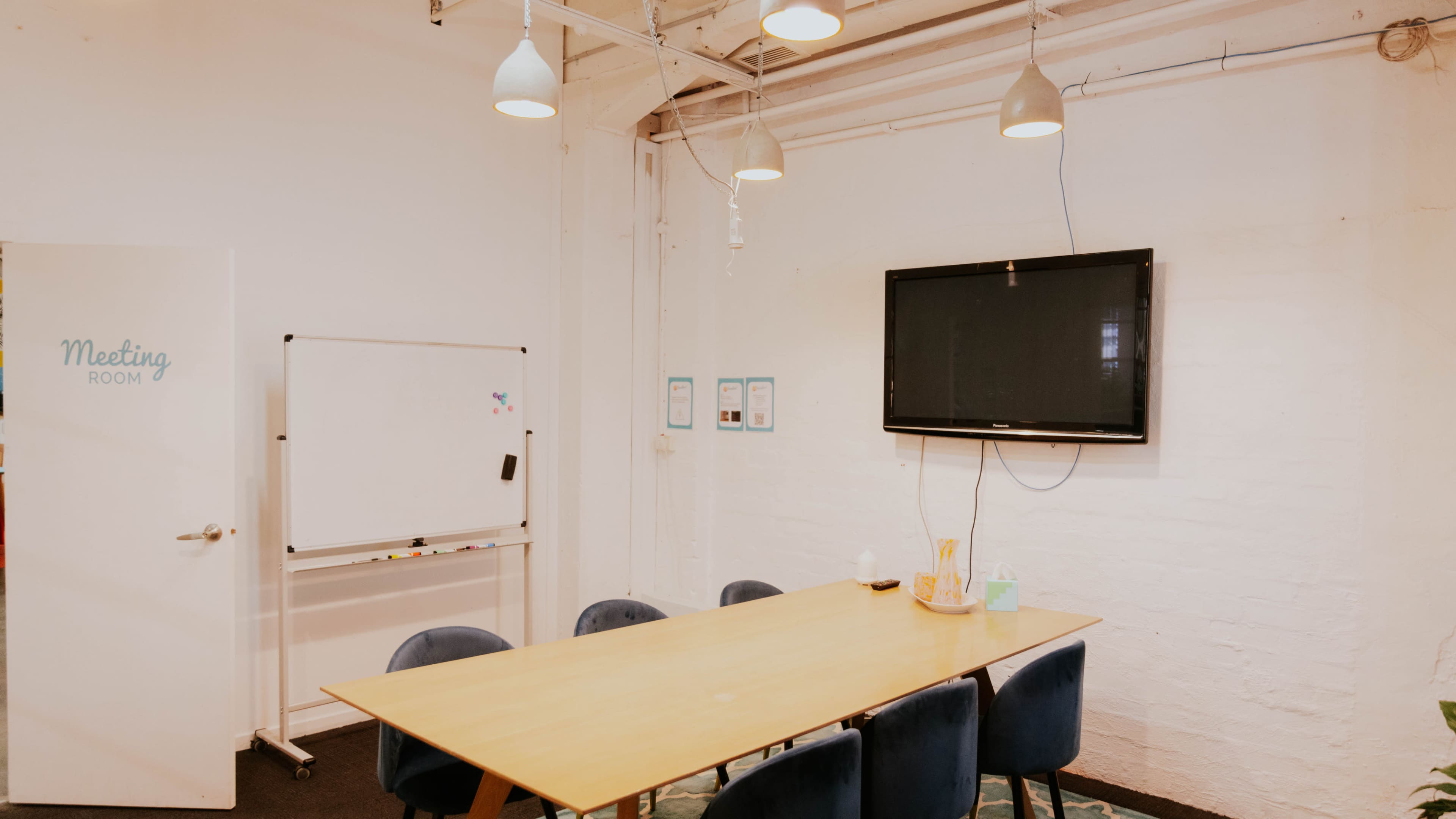 A meeting room features a long table surrounded by chairs, a whiteboard on wheels, and a mounted television on the wall.