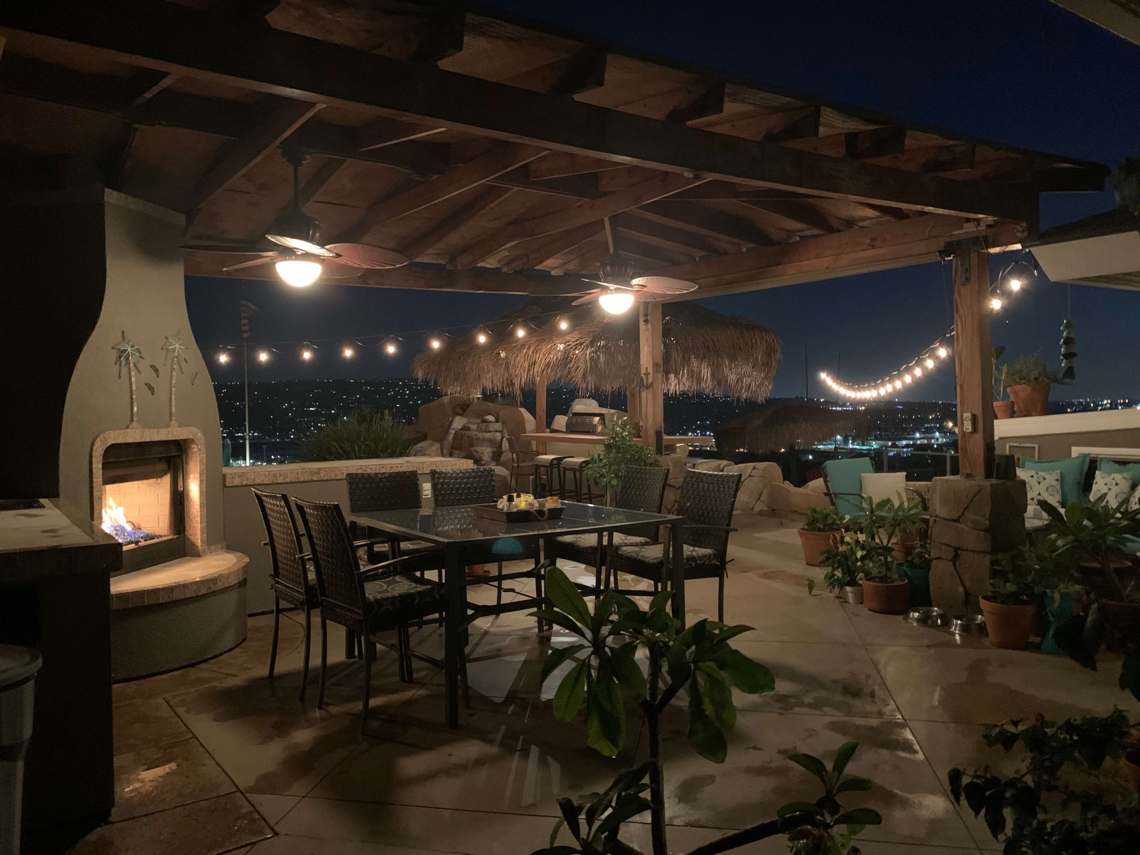 A patio at night with a dining table surrounded by chairs, a fire feature, and a thatched-roof lounge area, overlooking a city skyline adorned with lights.