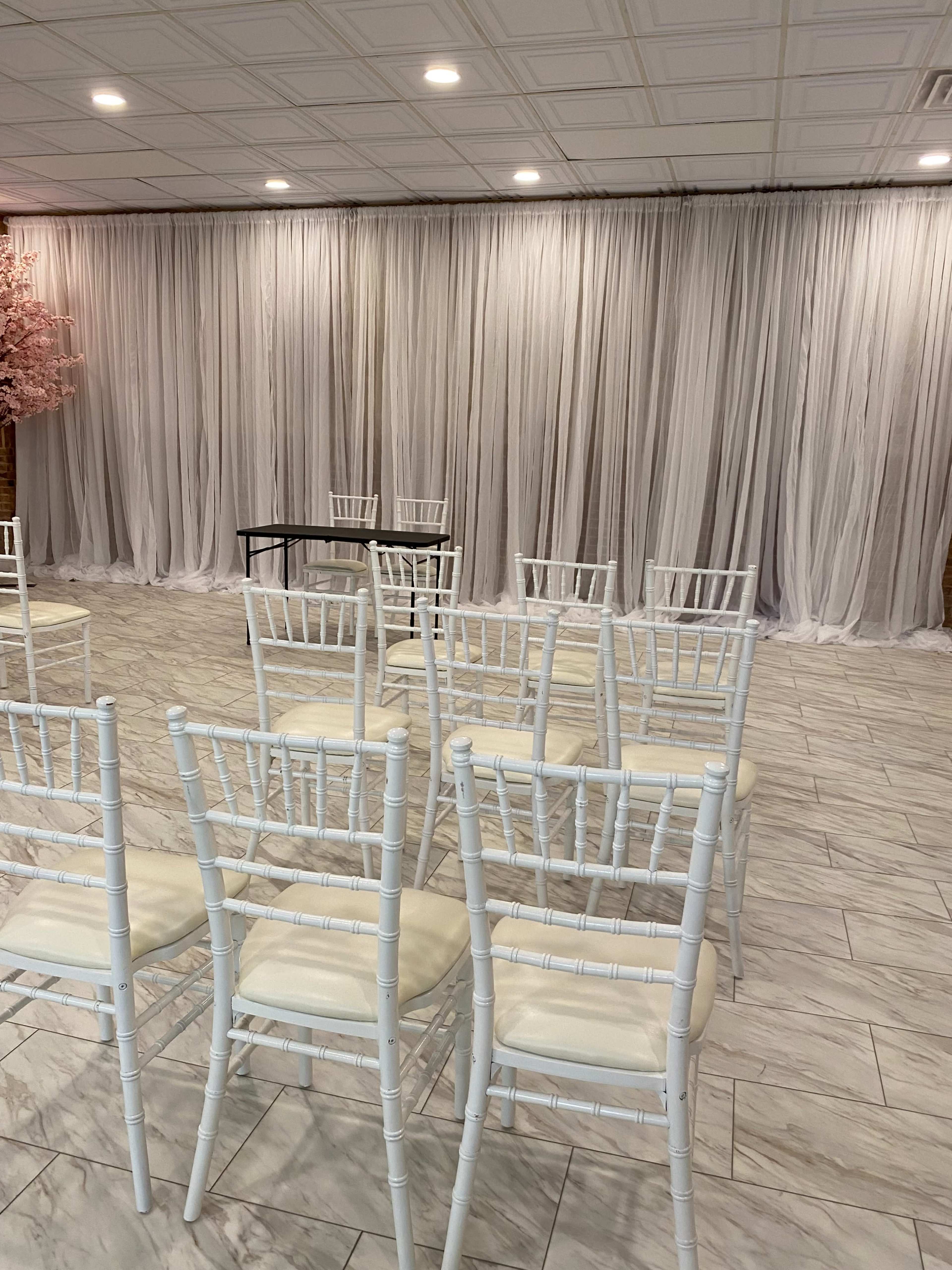 A set of white chairs is arranged in rows facing a table in front of a backdrop of sheer white curtains.