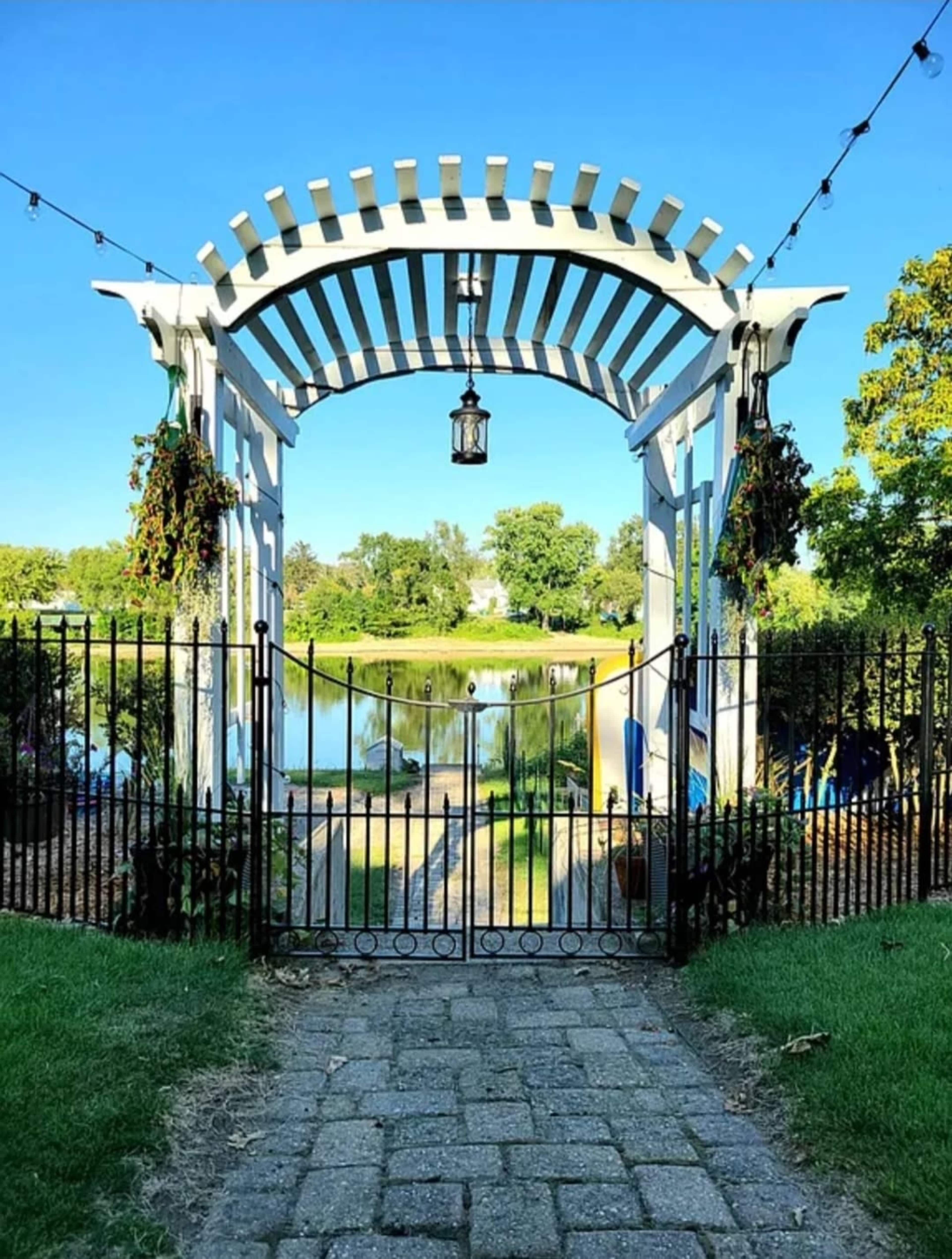 A white pergola with a lantern stands at the entrance of a path leading to a serene pond, surrounded by greenery and flower beds.