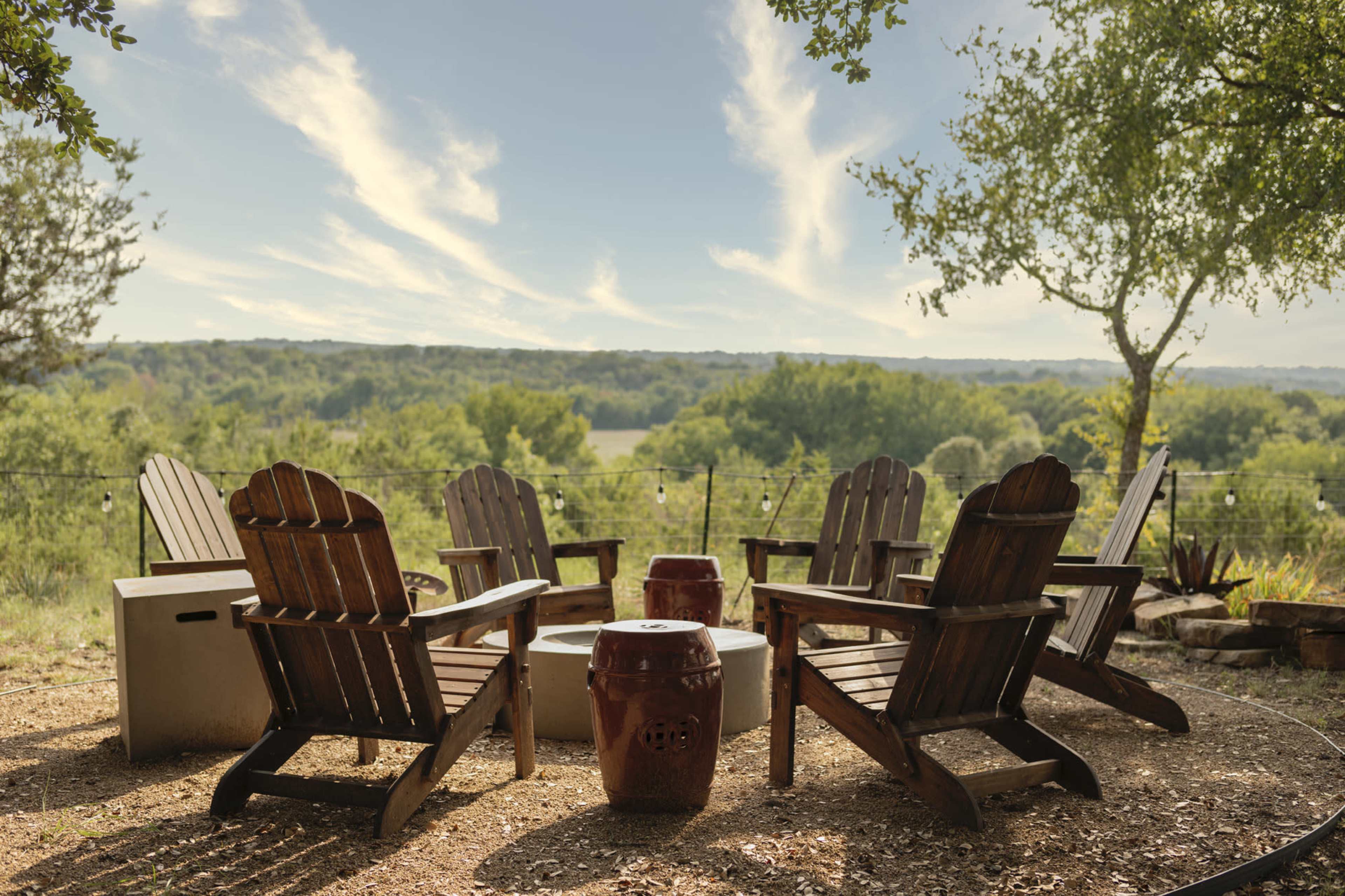 A circular arrangement of wooden Adirondack chairs surrounds a central stone fire pit, set against a backdrop of rolling green hills and a blue sky with wispy clouds.