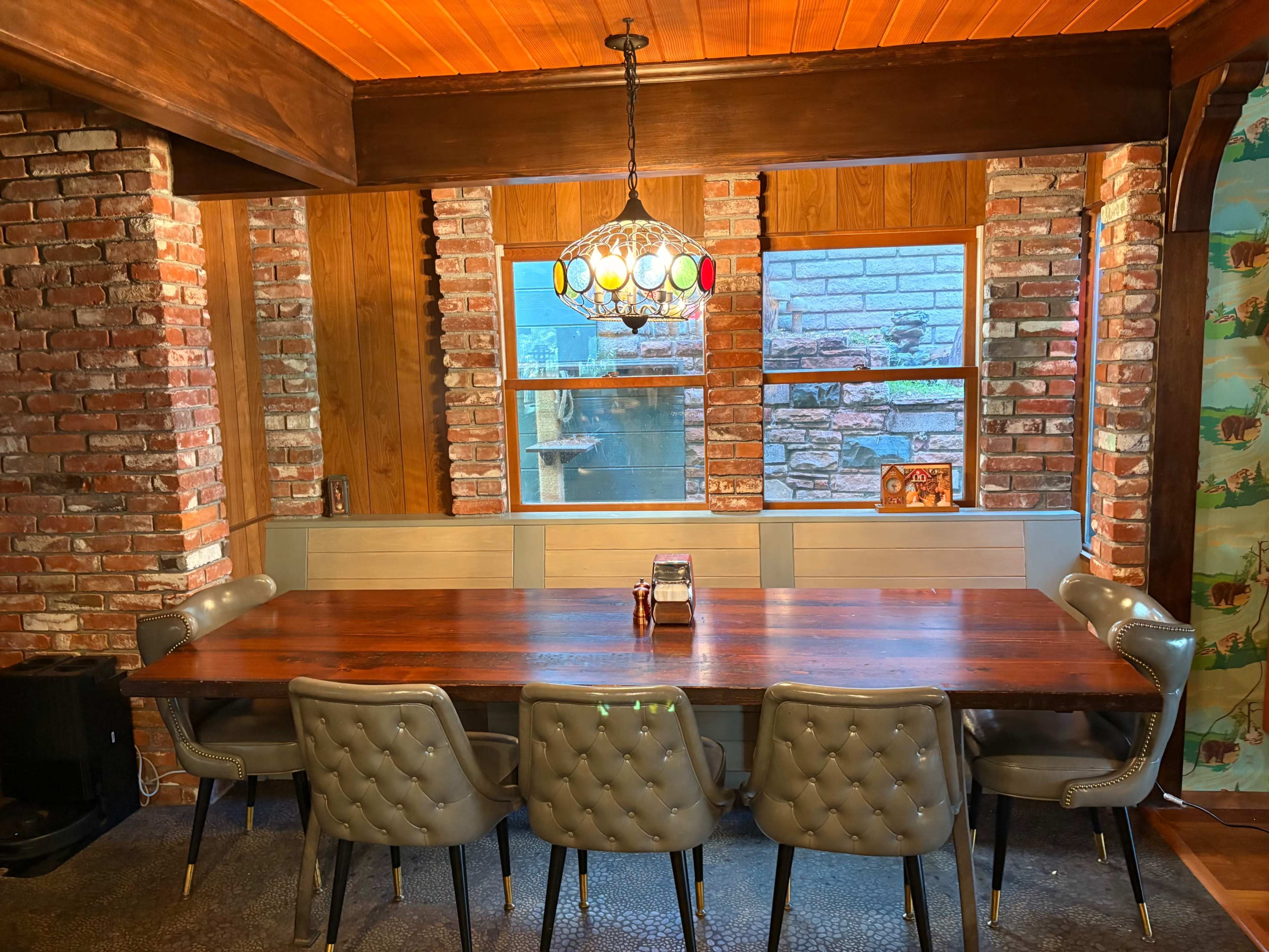 The image shows a rustic dining area with a large wooden table surrounded by tufted gray chairs, brick walls, and a decorative chandelier.