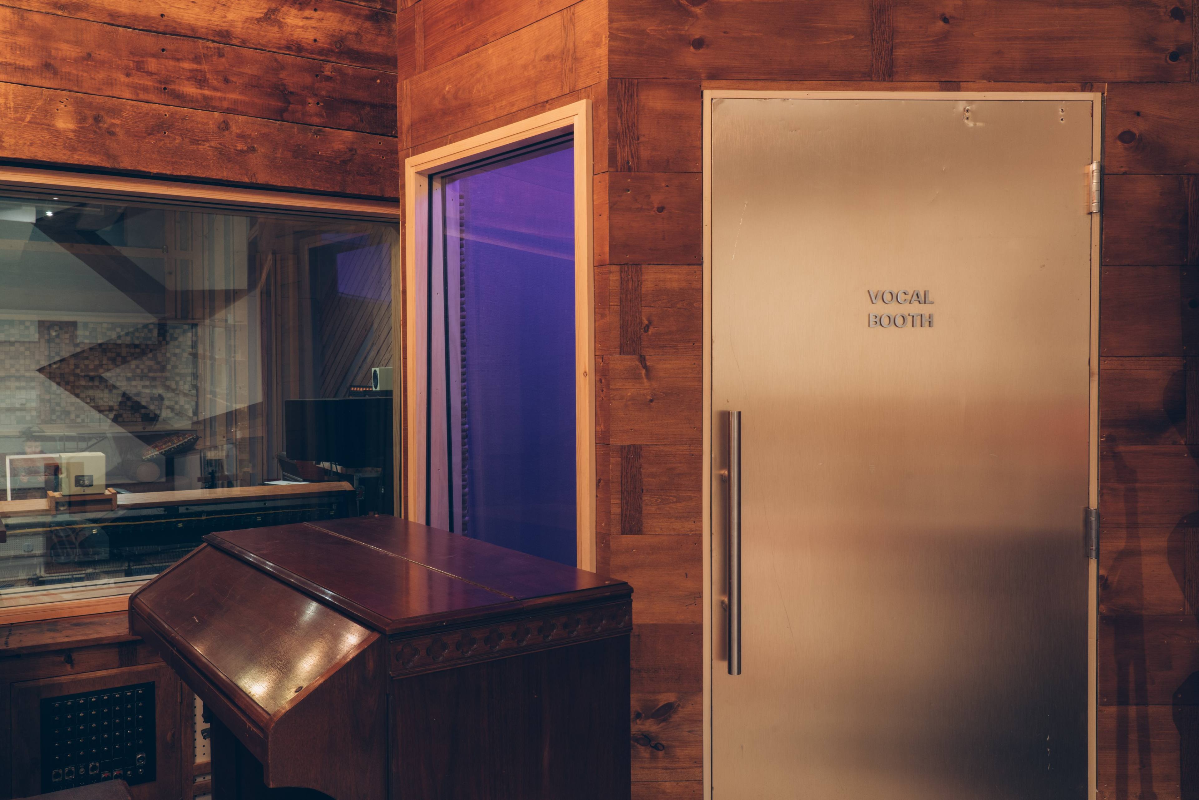 A wooden music studio features a glass-walled vocal booth with a metal door labeled "VOCAL BOOTH" and an antique wooden instrument nearby.