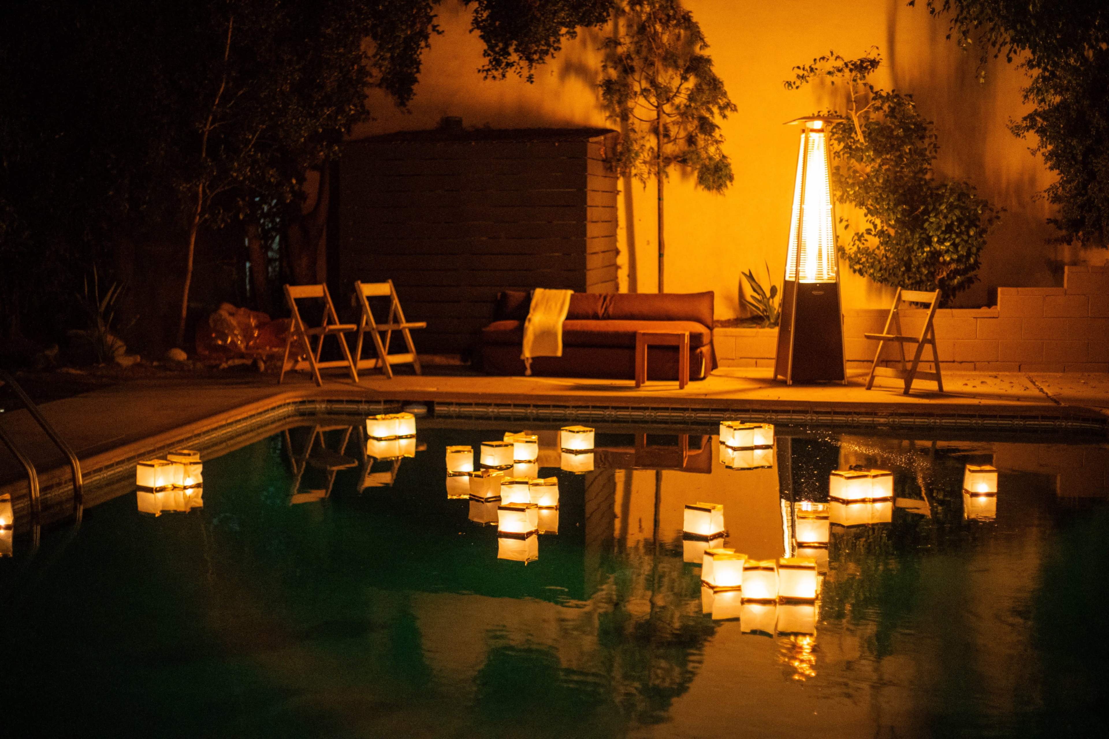 Lanterns float on the surface of a pool, illuminated by the warm glow of nearby lights and furniture in a private backyard setting.