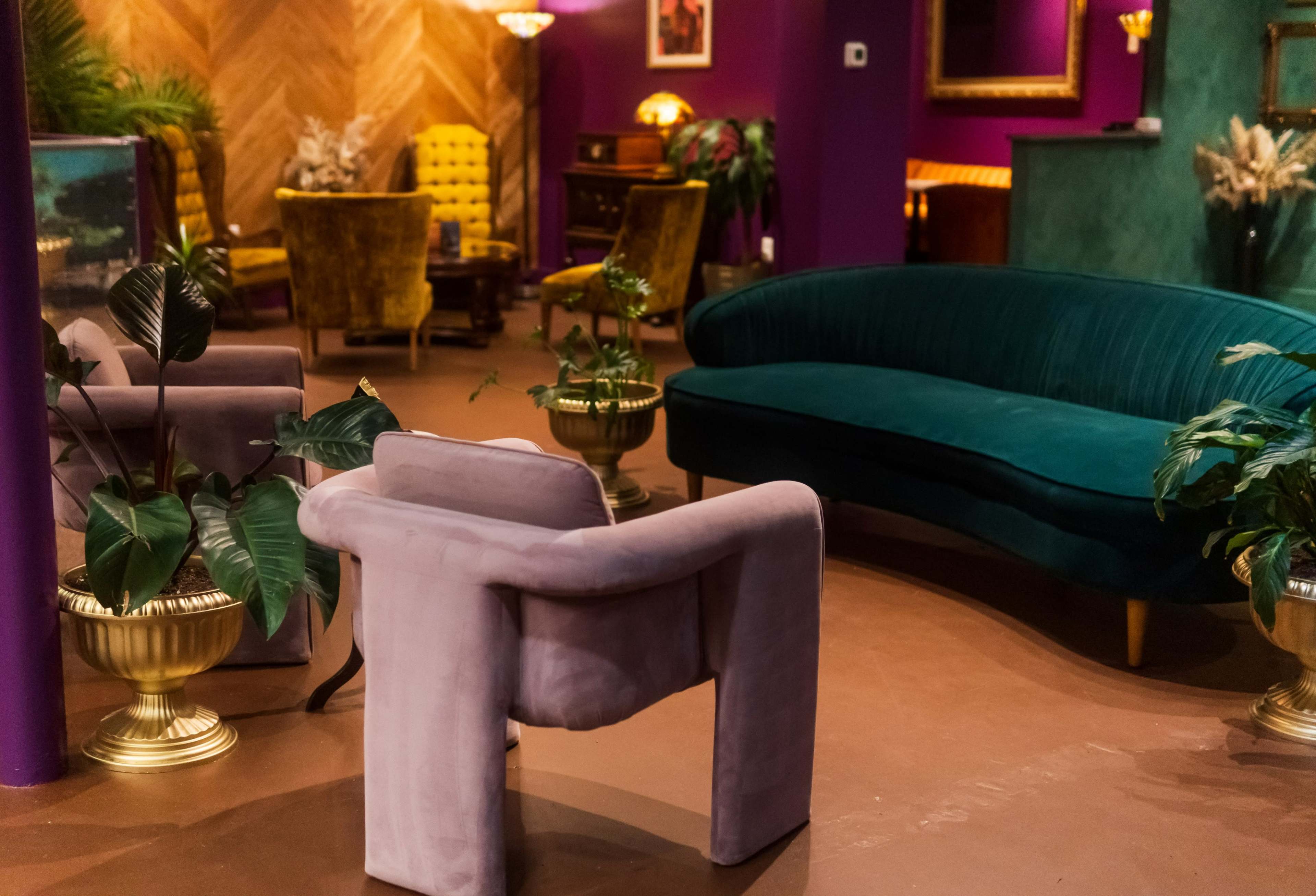 The image shows a cozy interior space featuring a green velvet couch, purple chairs, and potted plants arranged around a warmly decorated room with gold accents.