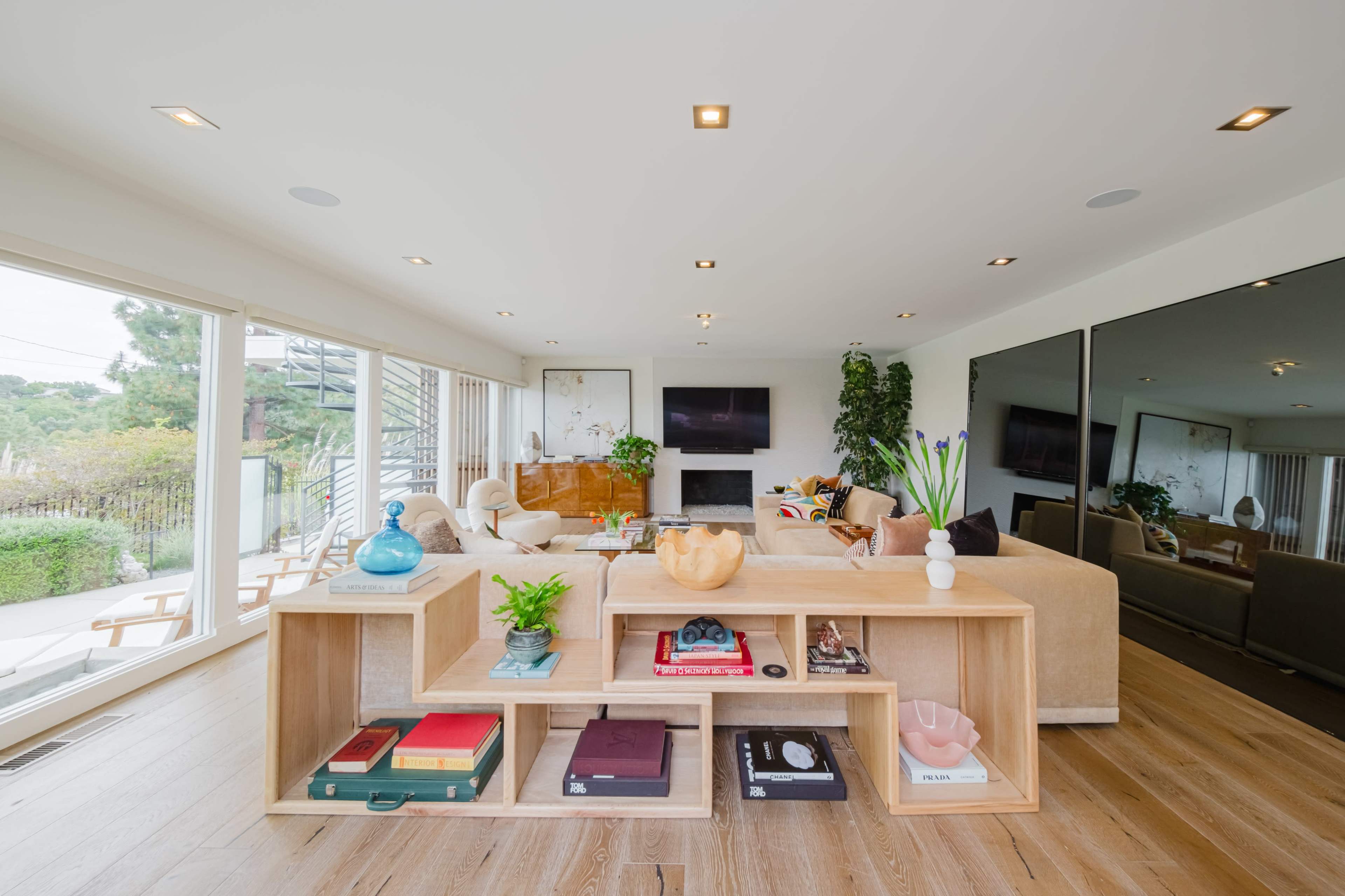 The image shows a modern living room with a light-colored sectional sofa, a wooden coffee table displaying books and decorative items, and large windows that provide a view of the outdoors.