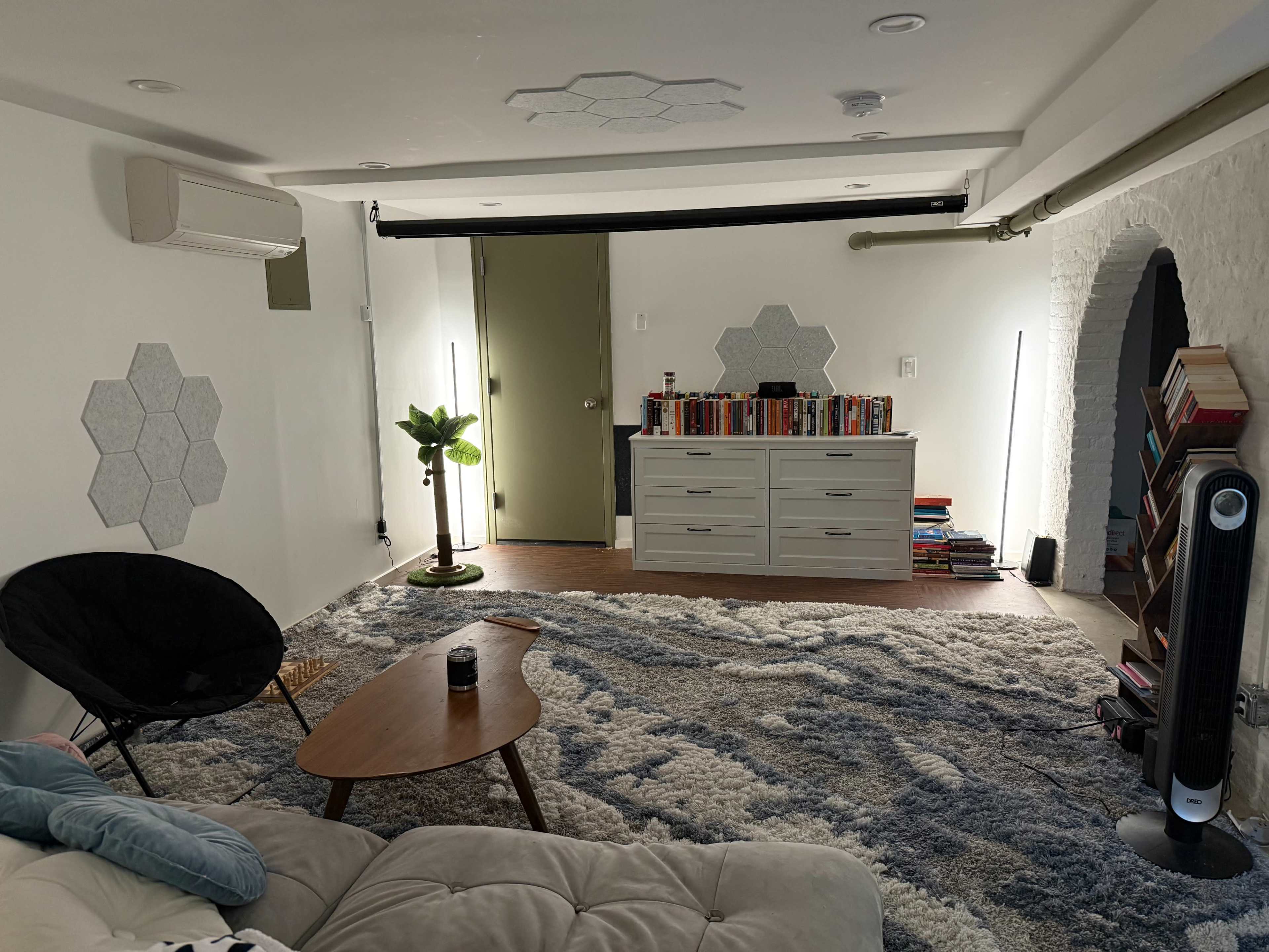 The image shows a modern basement room with a carpeted floor, a coffee table, a cozy chair, a set of bookshelves, and a decorative plant.