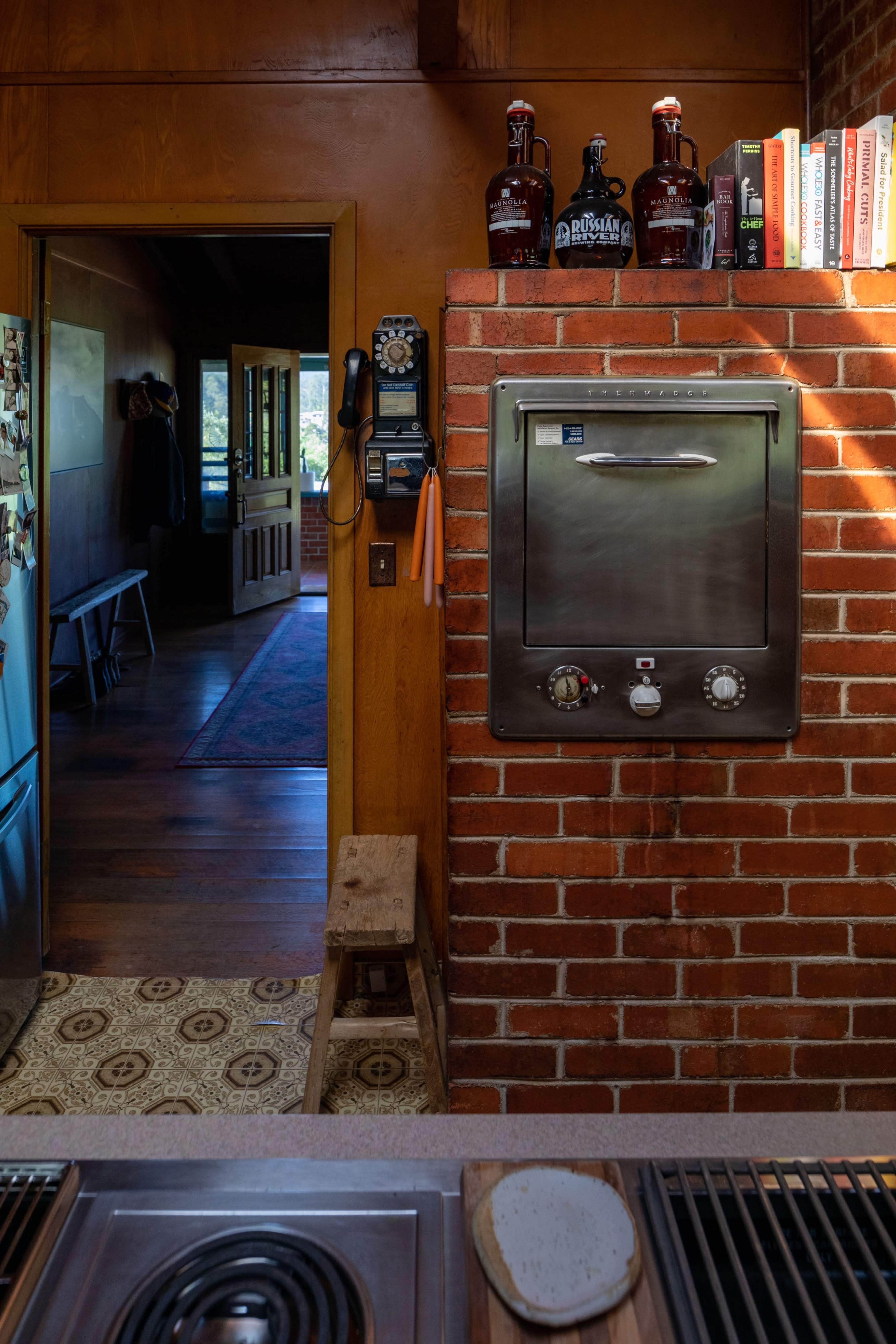 The image shows a kitchen with a brick wall, a metal oven, and a doorway leading to a hallway with wooden floors and a view of a door.
