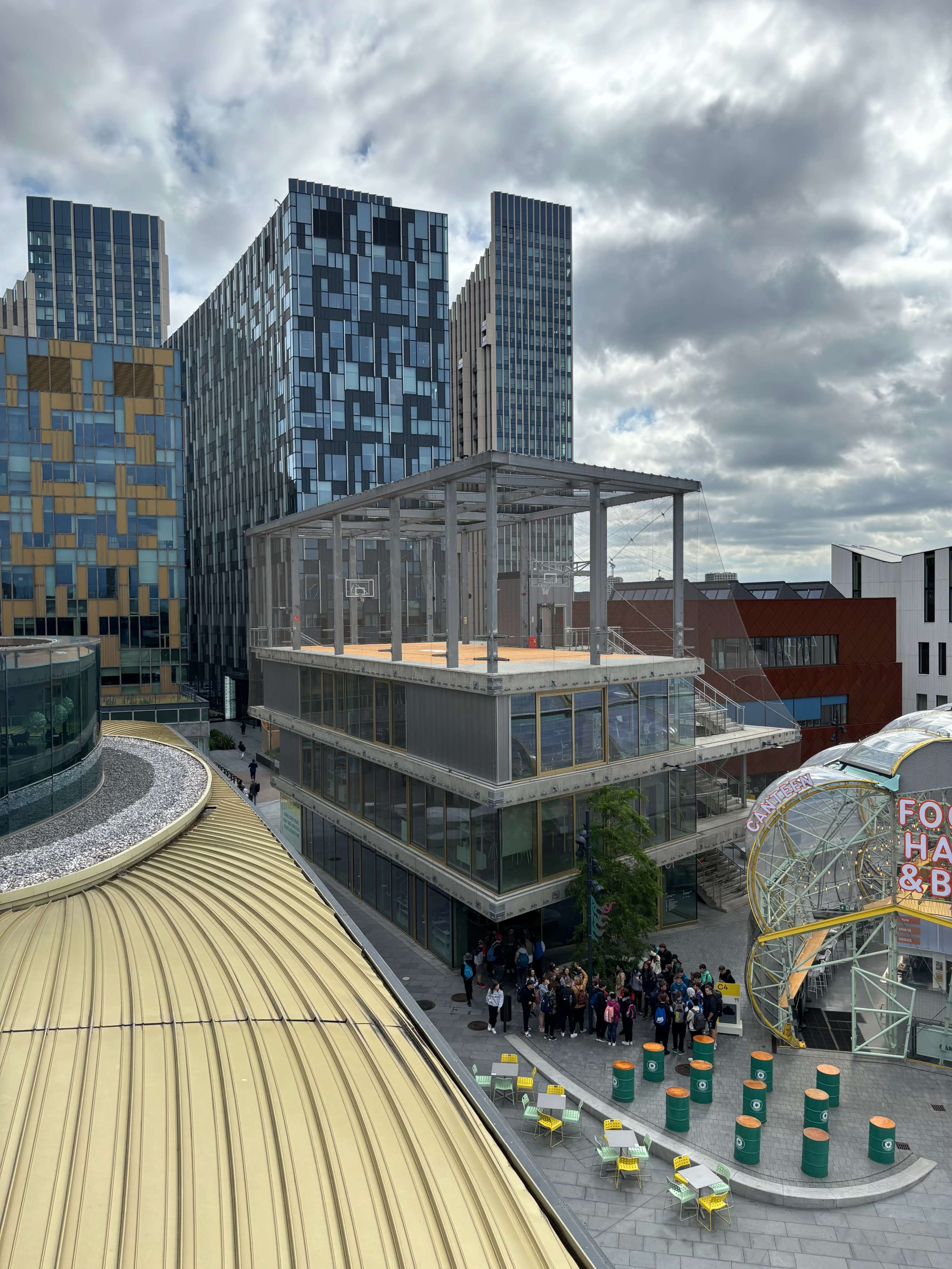 The image shows a modern urban area with several high-rise buildings, a partially constructed structure, and a group of people gathered near colorful cylindrical seating.