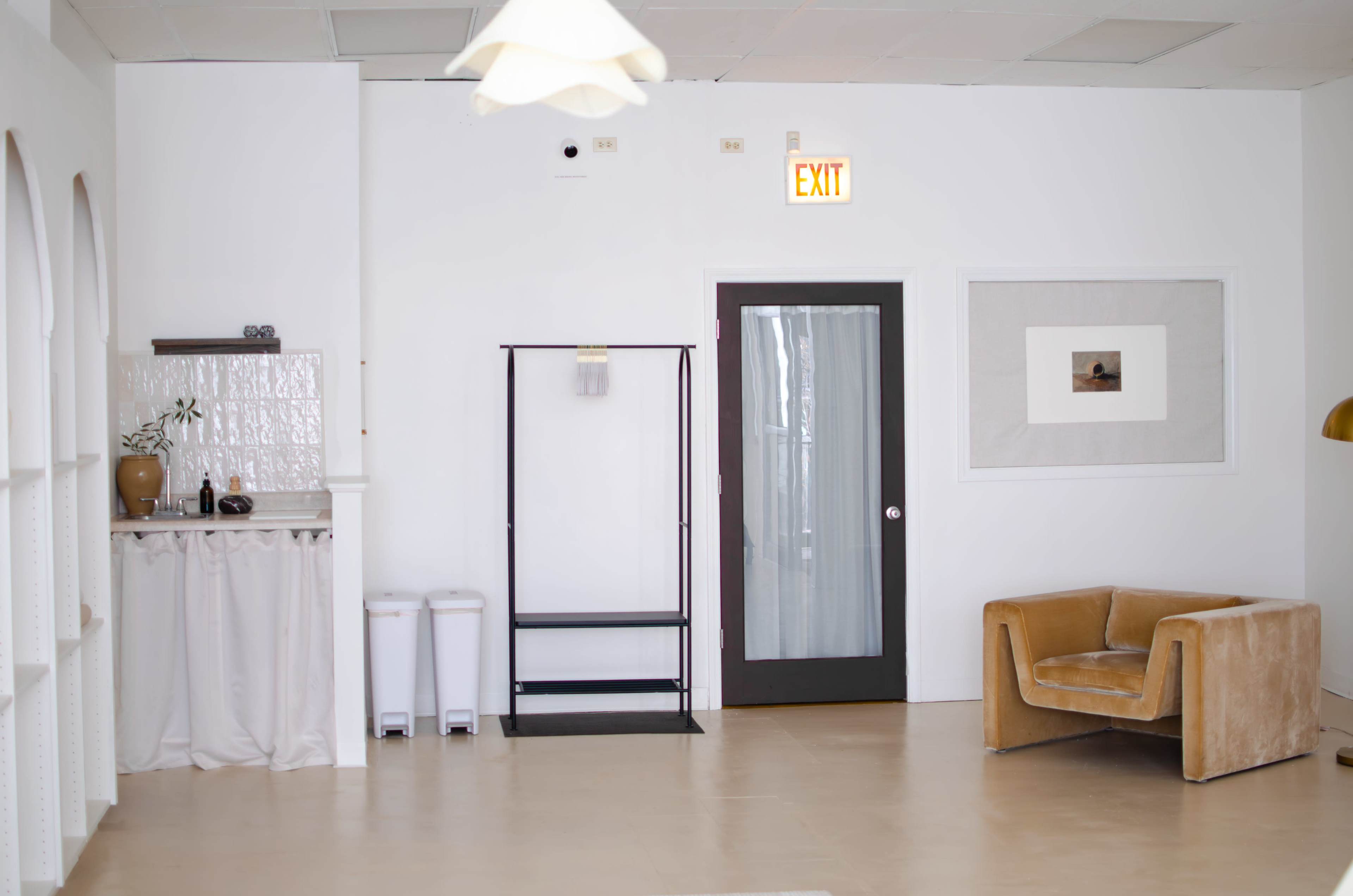 The image shows a minimalist interior space with a small kitchenette area, a coat rack, a door, and a stylish armchair against a white wall.