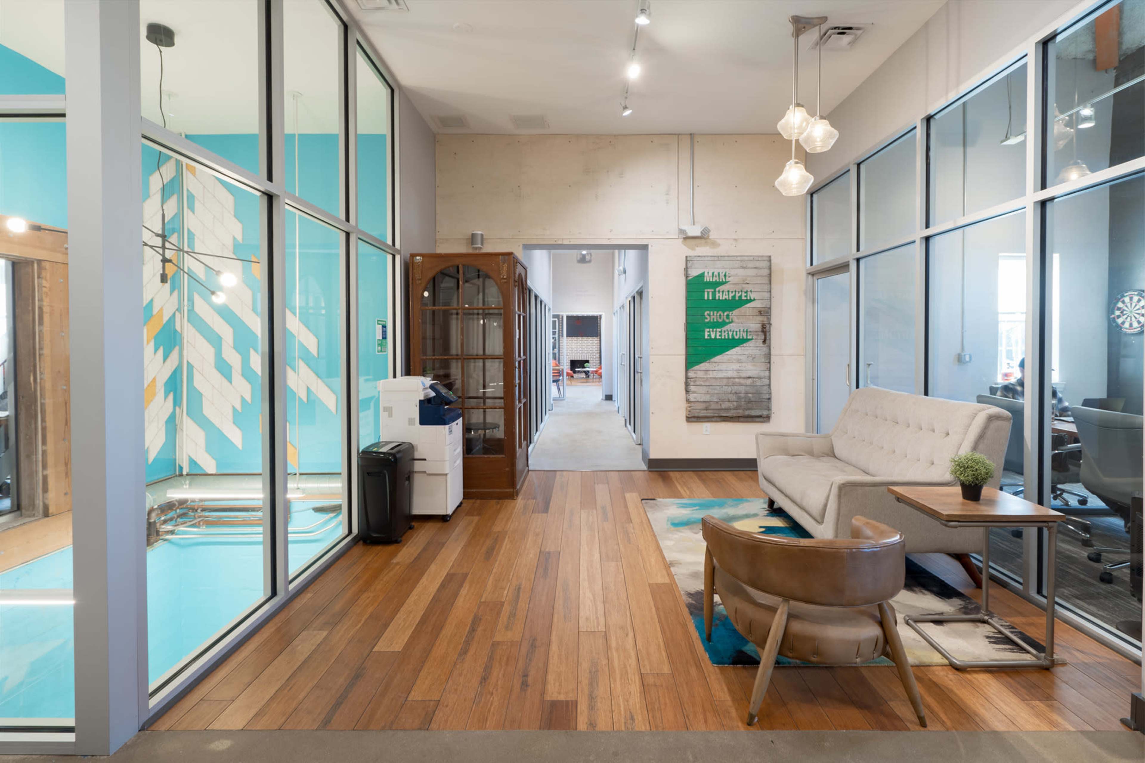 The image shows a modern office interior with wooden flooring, glass partitions, and a seating area featuring a sofa and a chair, along with a decorative wall and an open hallway.