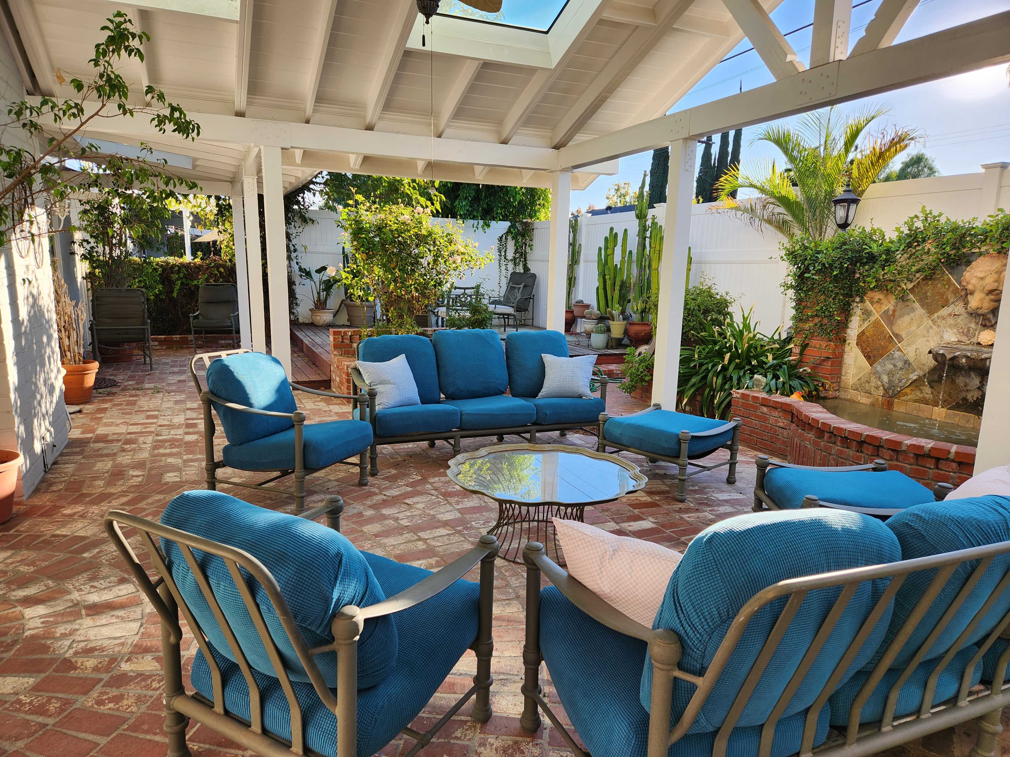 A patio area with blue upholstered seating arranged around a glass table, surrounded by greenery and a water fountain in the background.