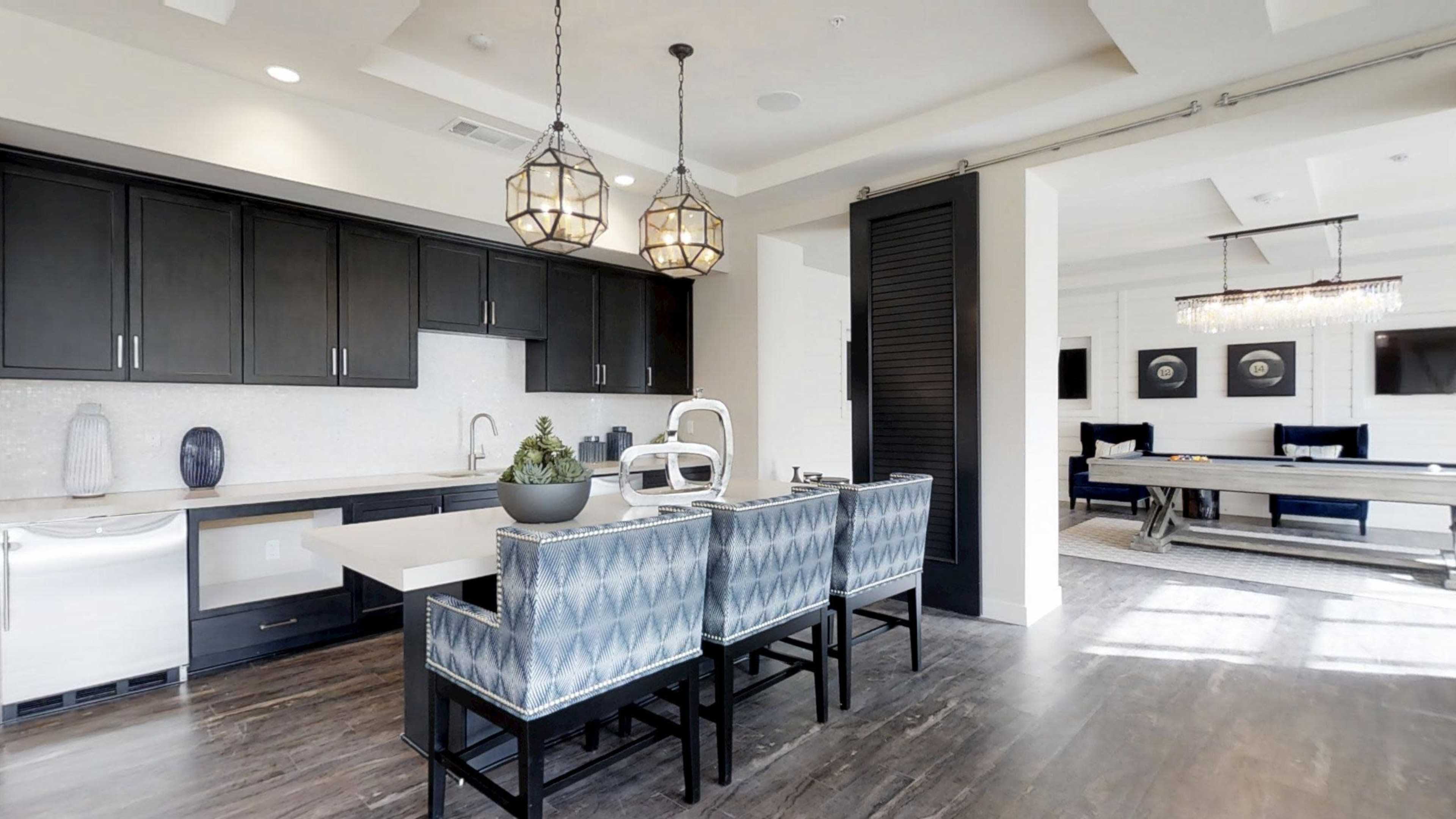 The image depicts a modern kitchen and dining area with a large island, bar stools, dark cabinetry, and a bright seating area featuring a long table and decorative art.