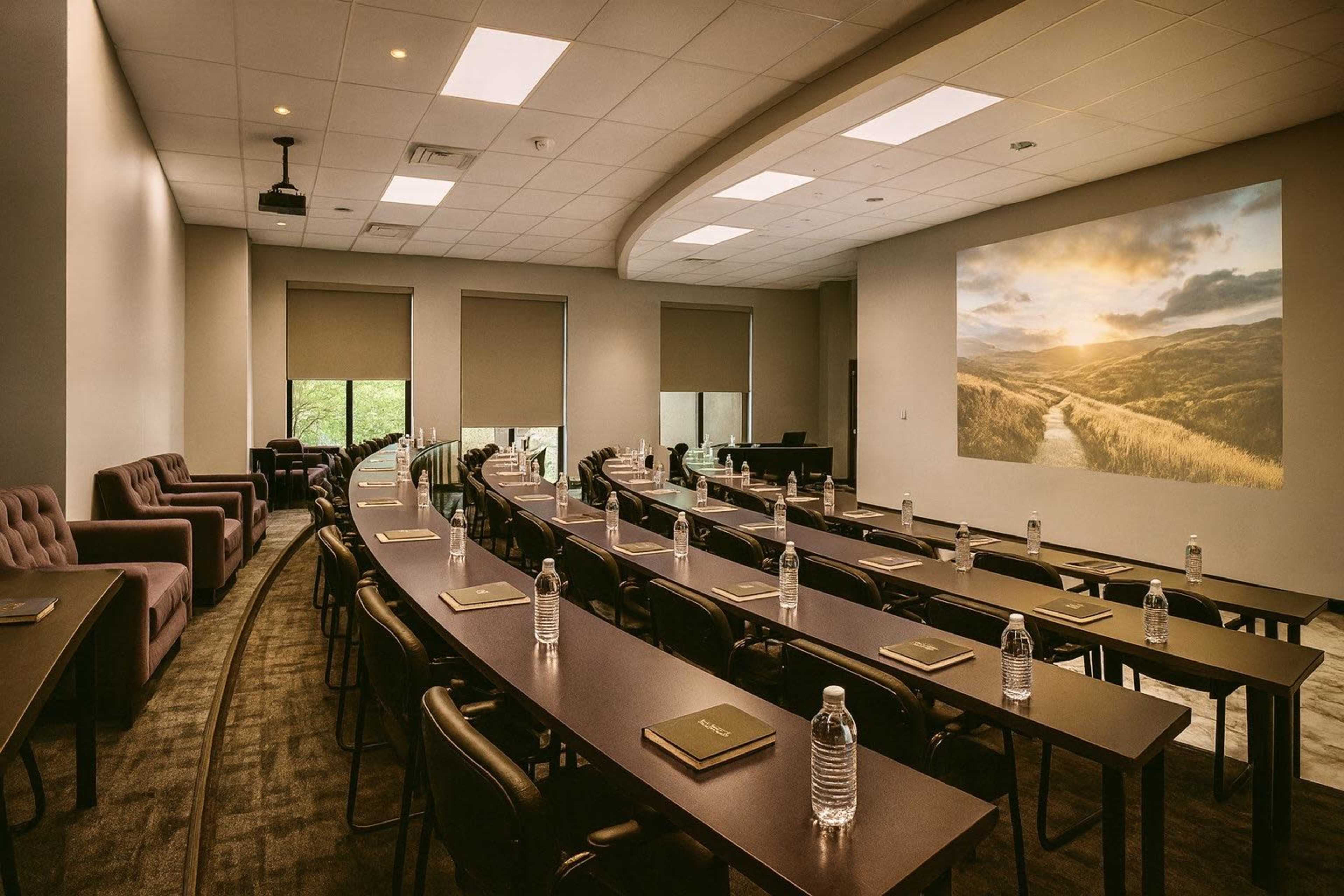 The image shows a conference room arranged with long tables and chairs, featuring a projector screen displaying a scenic landscape.
