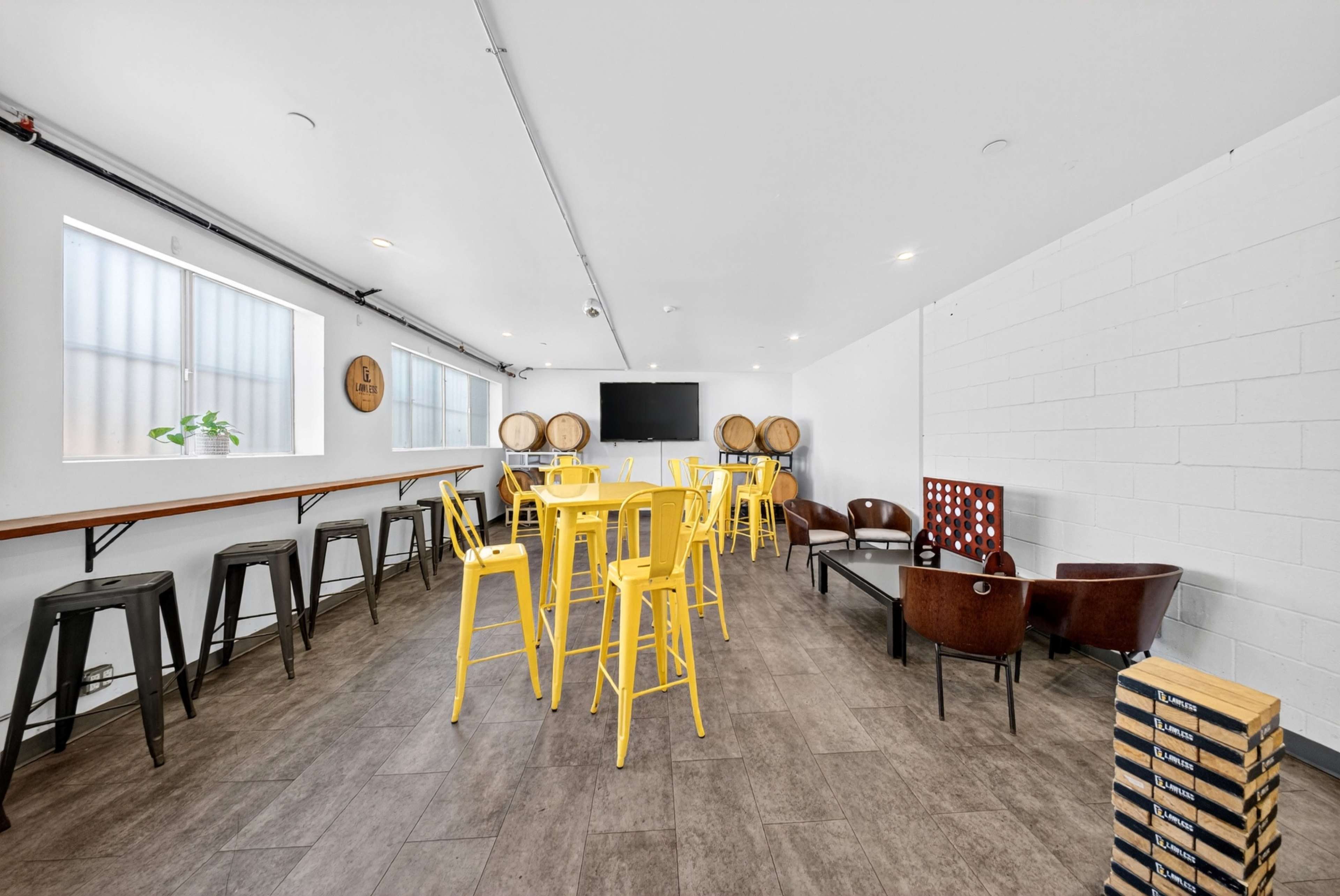 The image shows a spacious, modern room with yellow chairs, a long bar table, and a seating area, featuring a television and barrels against the white walls.
