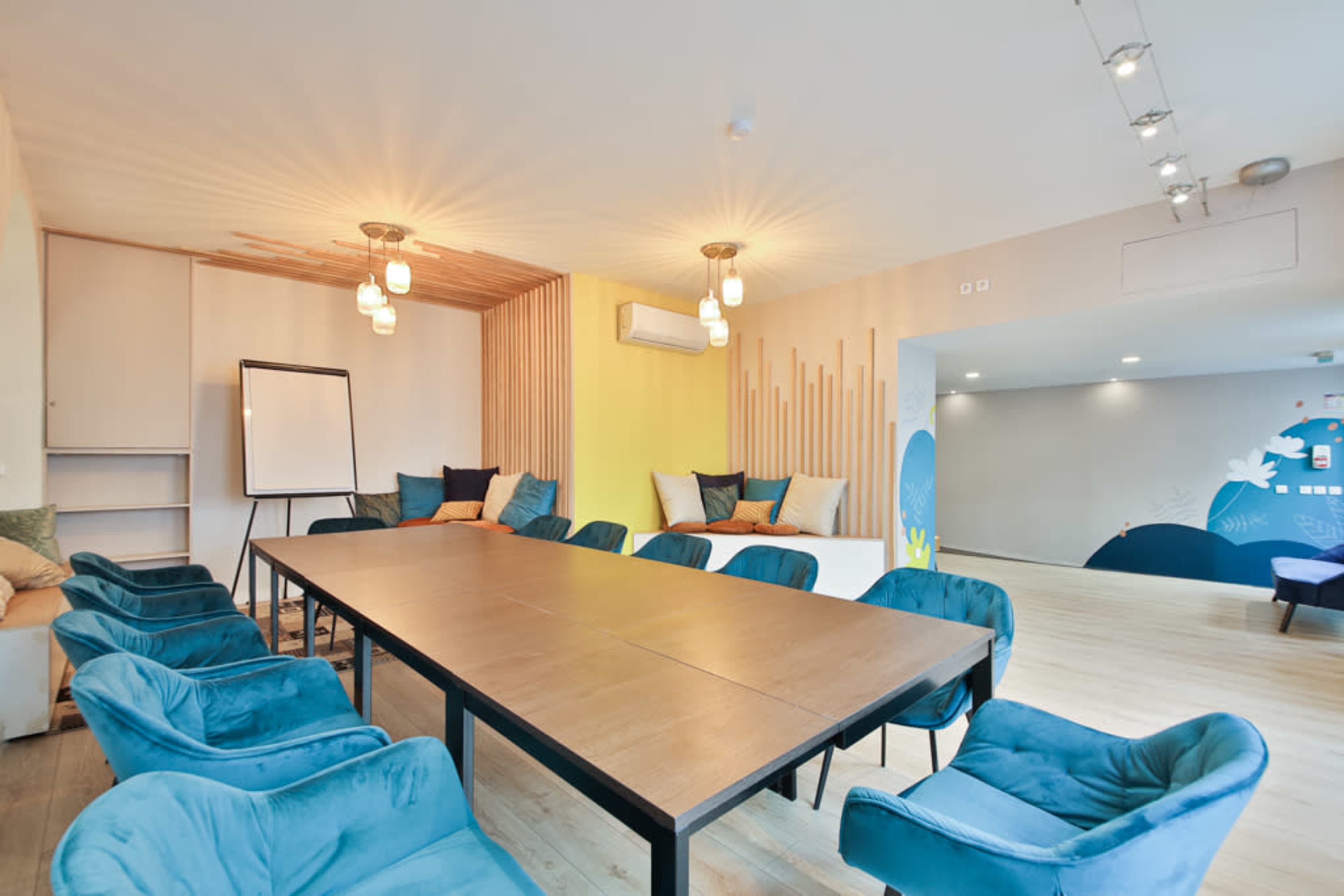A modern conference room features a long wooden table surrounded by teal chairs, with a whiteboard and cozy seating areas against the walls.