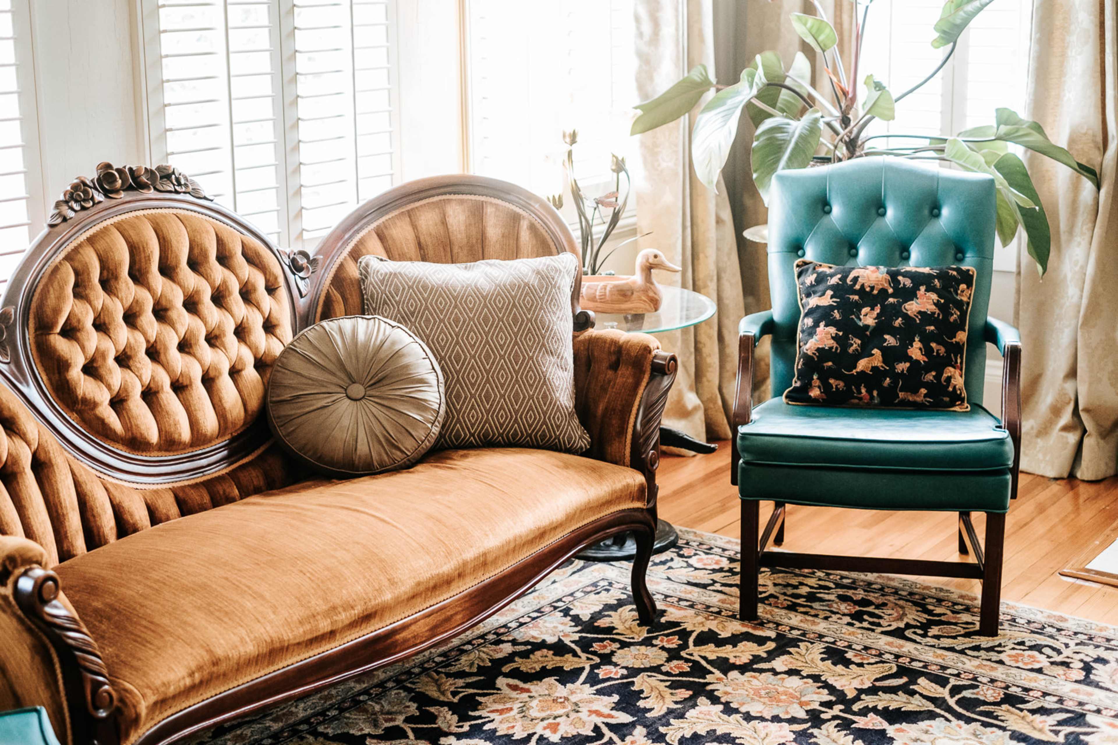 A vintage living room setup featuring a brown upholstered sofa with decorative cushions and a teal upholstered chair beside a small round table and a potted plant.